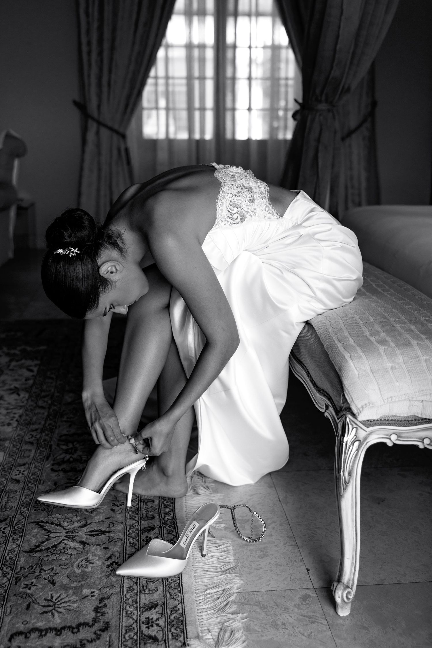 A black and white photo of someone in a white dress fastening their high heel shoe while sitting on an elegant chair.
