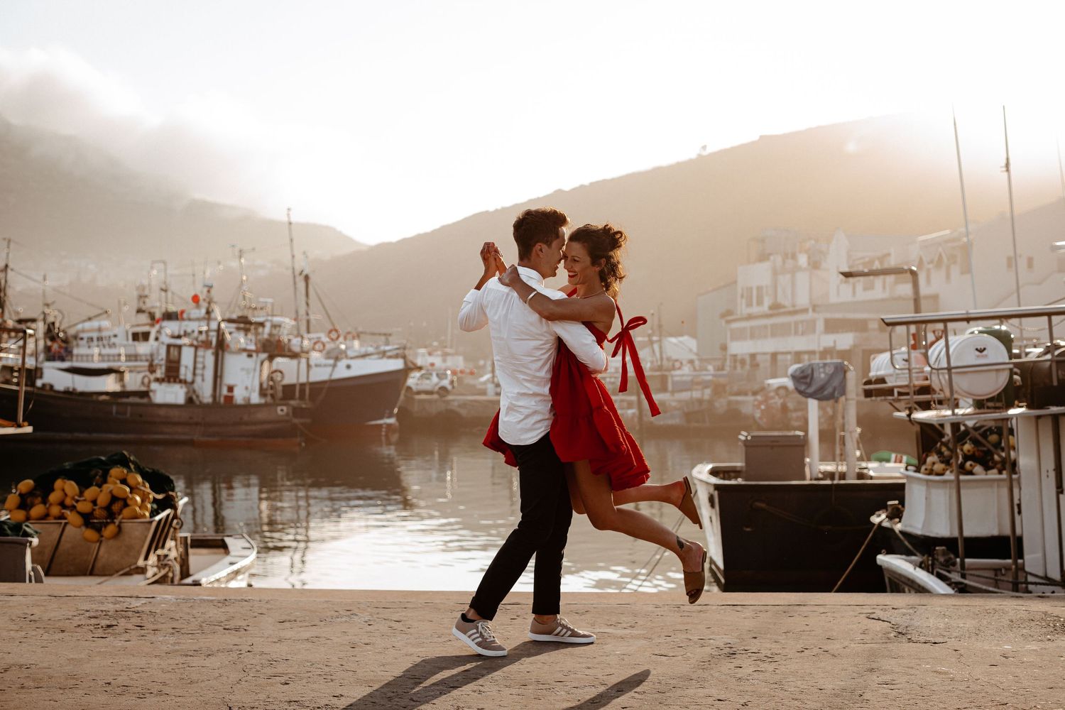 A couple dances romantically at sunset on a dock with boats and mountains in the background.