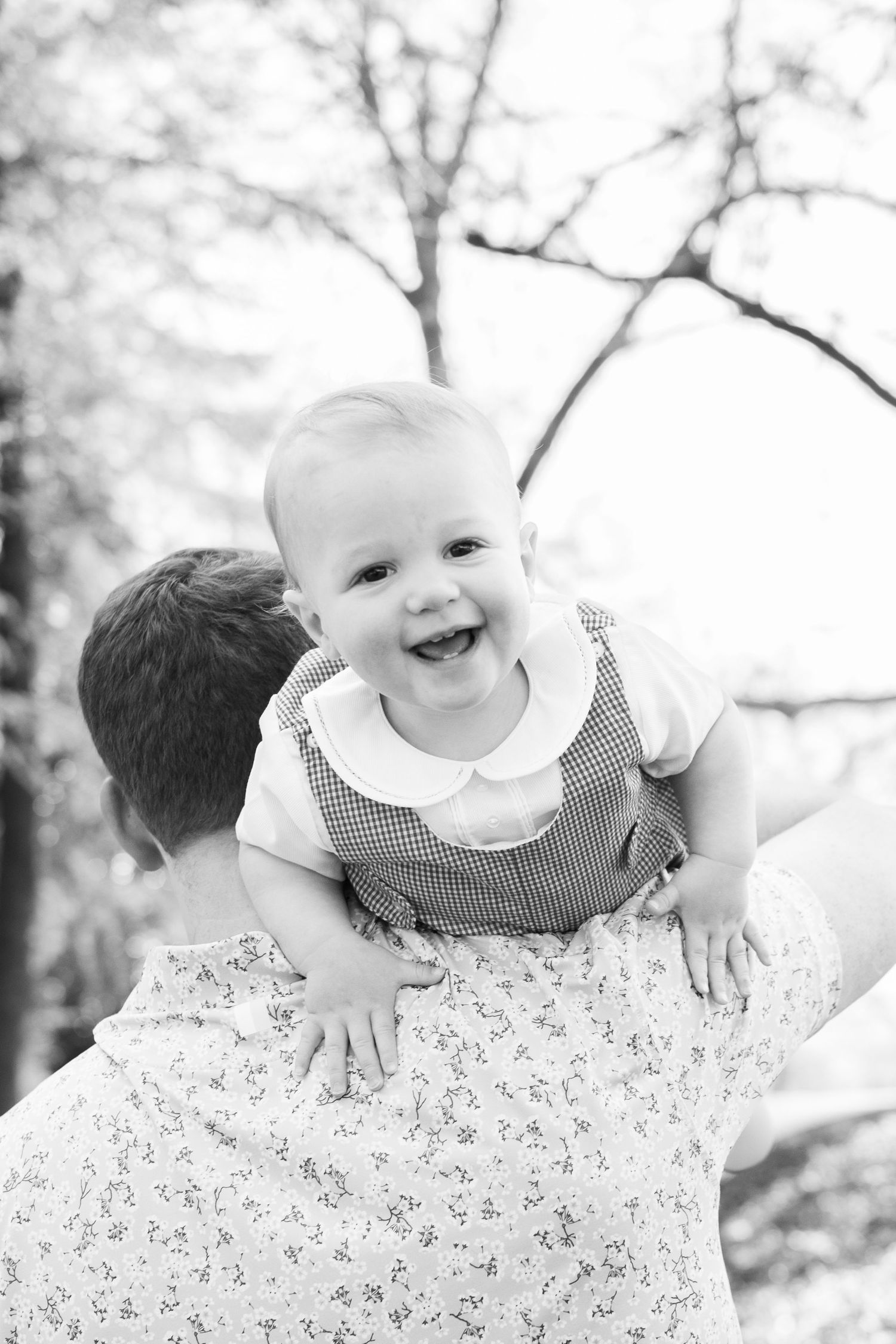 Black and white photo of a laughing baby being held up by an adult outdoors near trees.