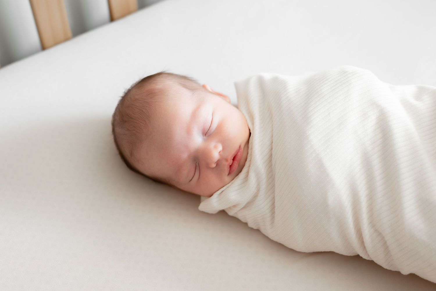 Peaceful newborn baby swaddled in white blanket sleeping in a crib.