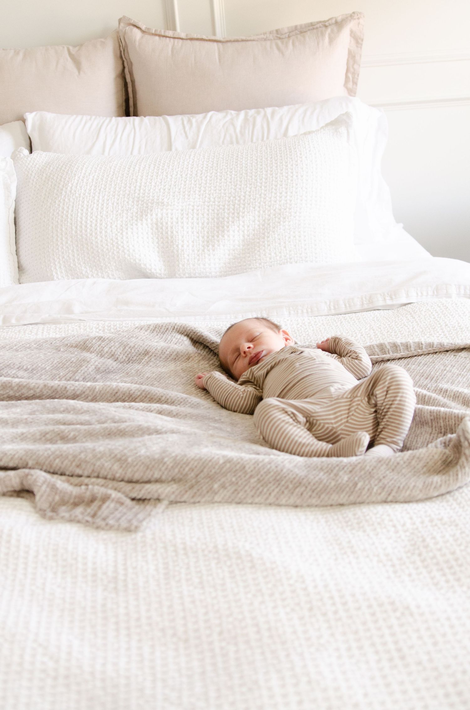 Baby in neutral colored onesie sleeping peacefully on white bedding in soft natural light.
