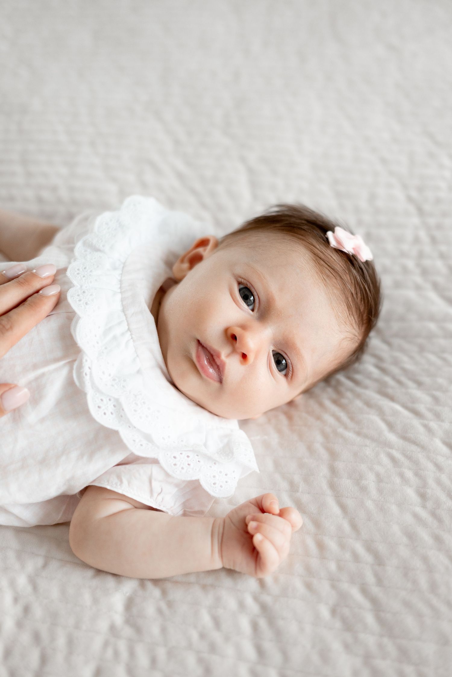 Newborn baby girl in white ruffle dress with pink bow hair clip lying on white quilted blanket.