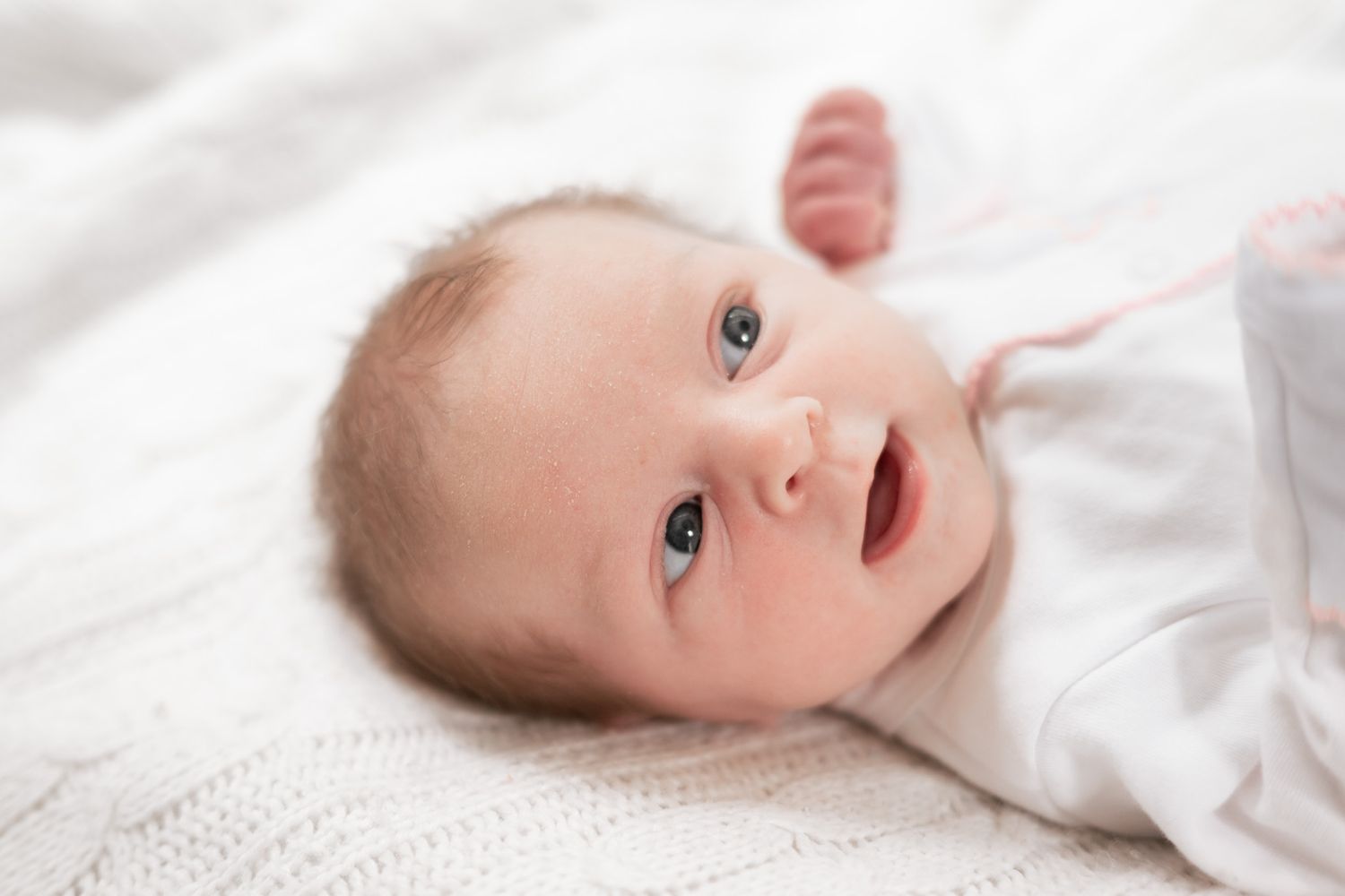 A newborn baby with blue eyes lying on a white knit blanket, wearing a white onesie, looking up curiously.