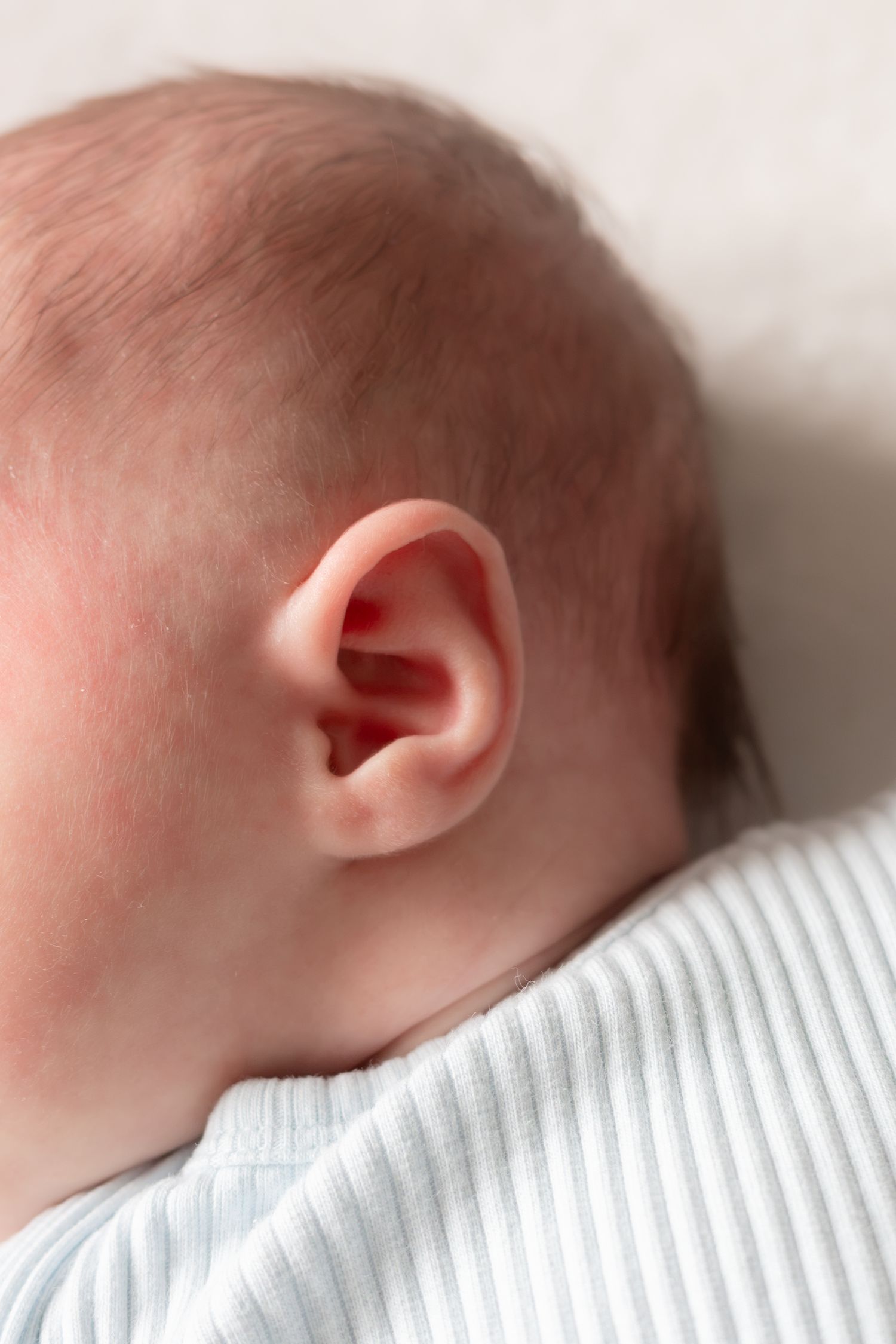 Close-up of a newborn baby's ear and head, with fine hair and soft skin, wearing a light blue striped outfit.
