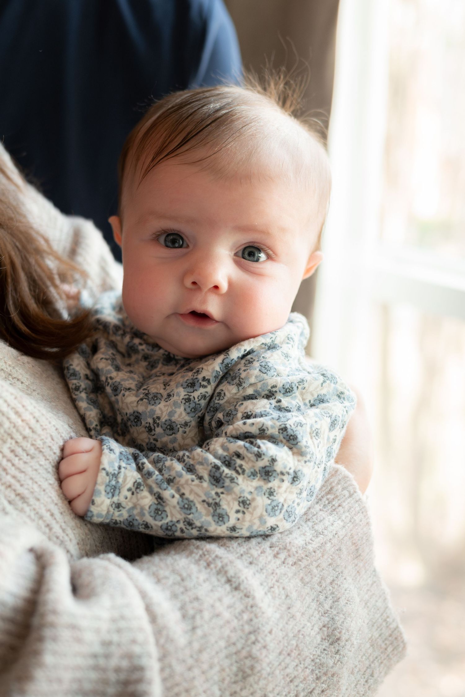 Baby in a floral sweater resting against a knit sweater by a window during a lifestyle newborn photography session in Charlotte, NC.