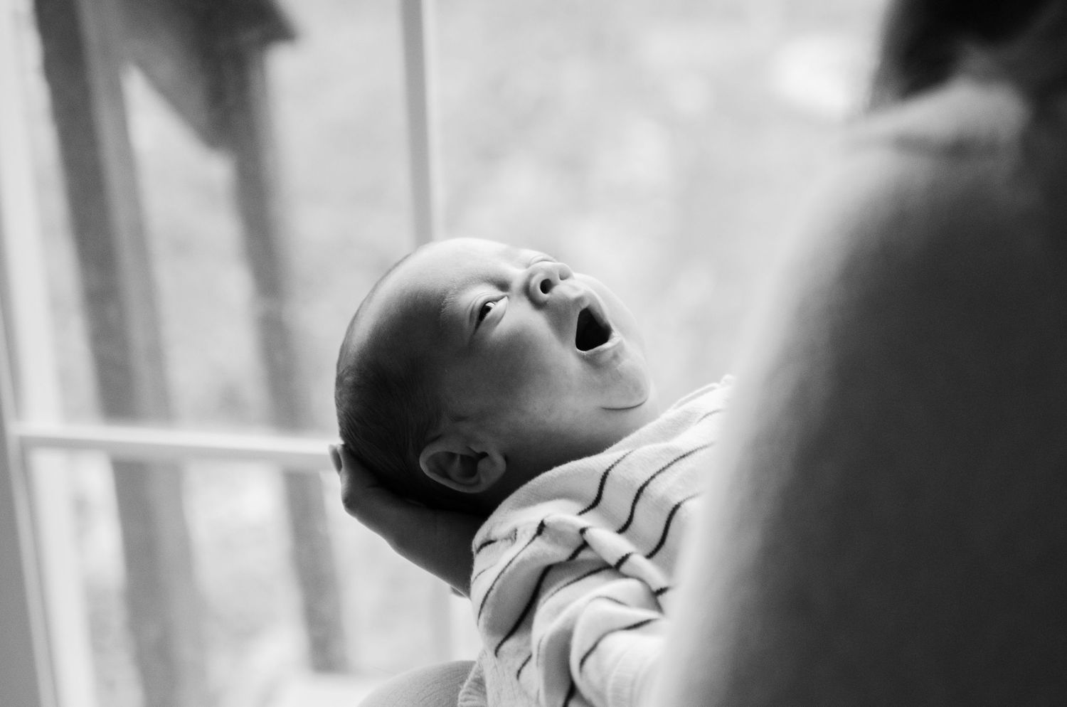 Black and white image of a baby yawning while being held near a window during a lifestyle newborn photography session in Charlotte, NC.