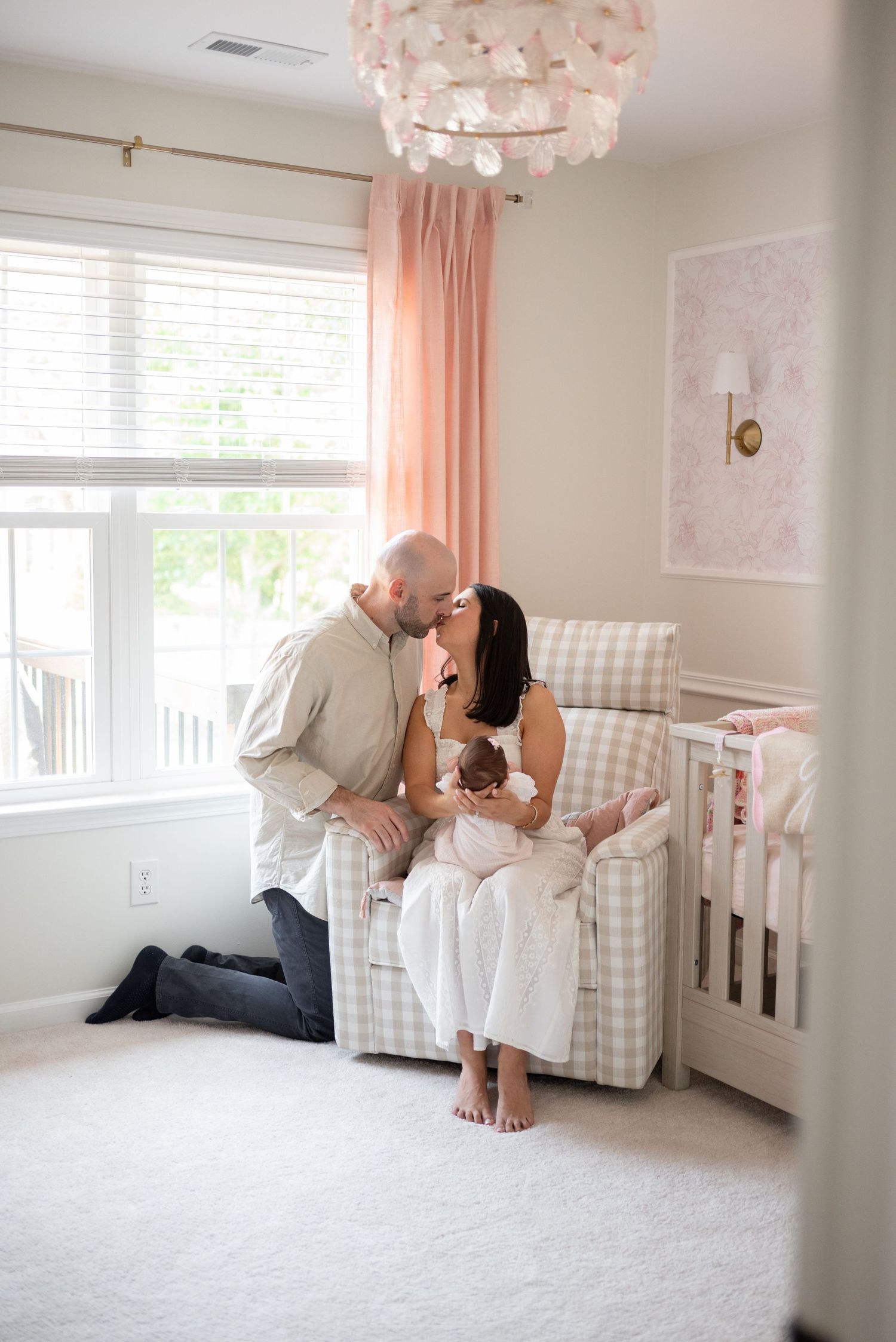 New parents sharing a tender kiss while holding their newborn in a soft pink nursery with a glider chair and crib.