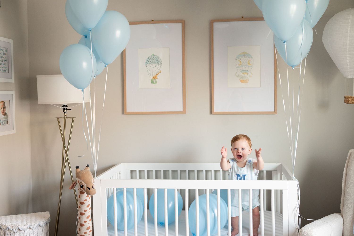 Happy baby standing in white crib surrounded by blue balloons in a decorated nursery room with framed animal prints.