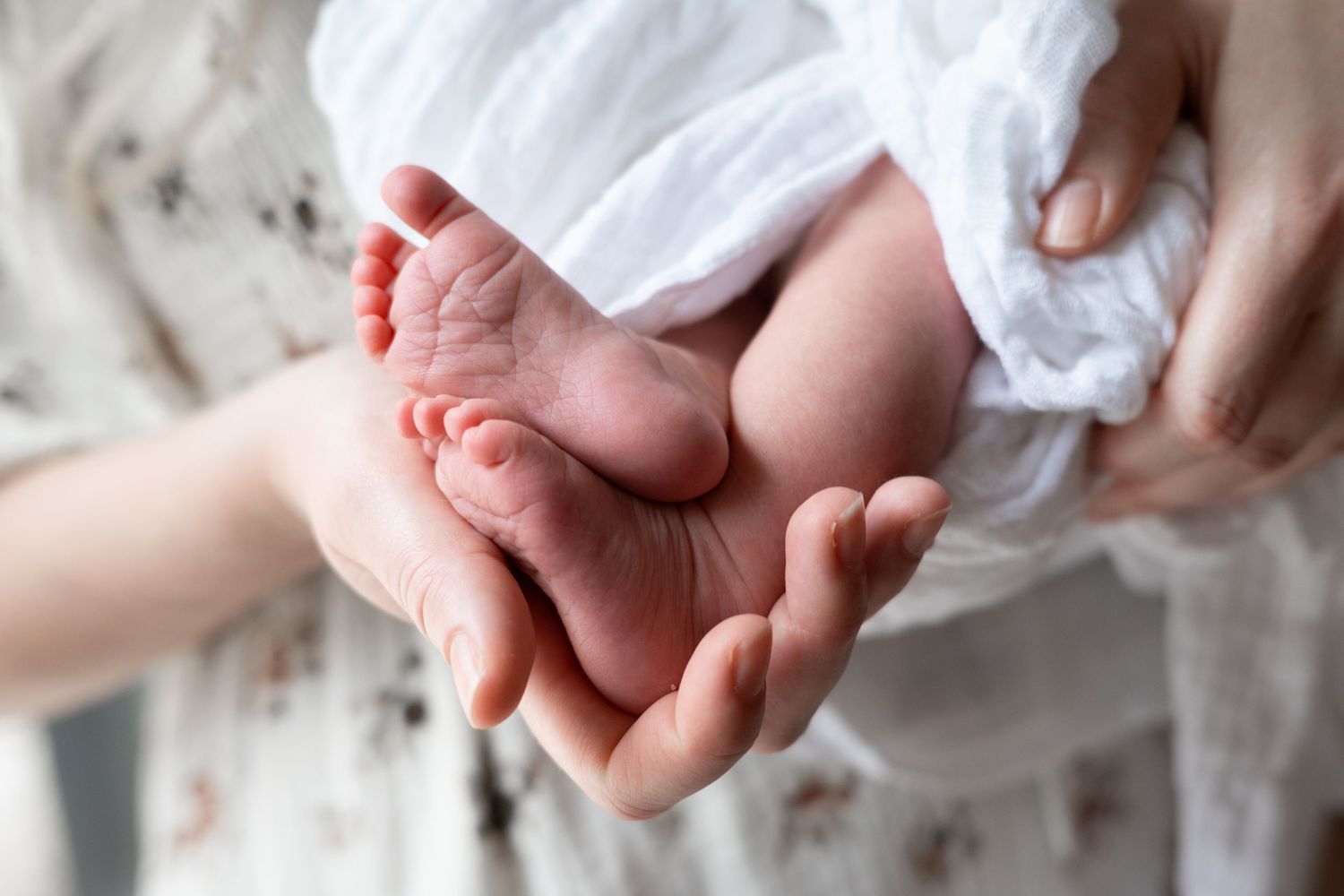 Close-up of a newborn baby's tiny feet cradled gently in a parent's hands, wrapped in a white swaddle.