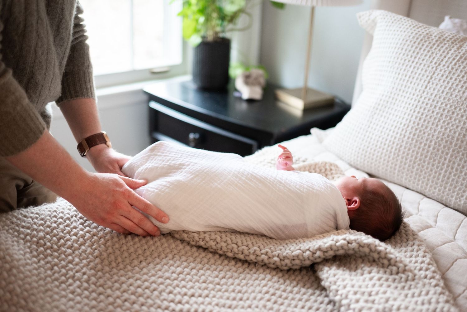 Parent swaddling a newborn baby in white muslin wrap on a bed with neutral bedding.