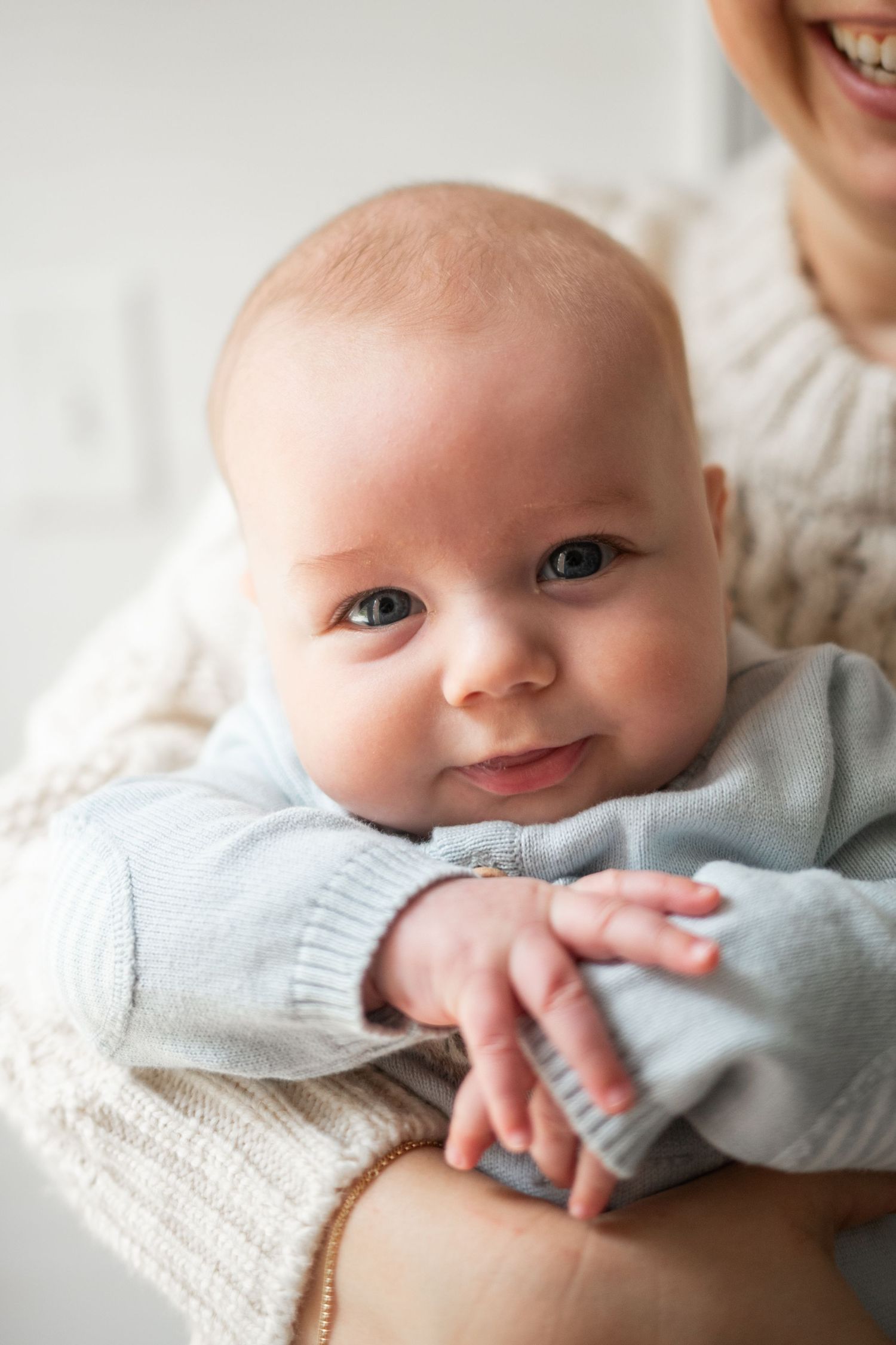 Smiling baby in a light blue sweater lying on cream fabric during a lifestyle newborn photography session in Charlotte, NC.
