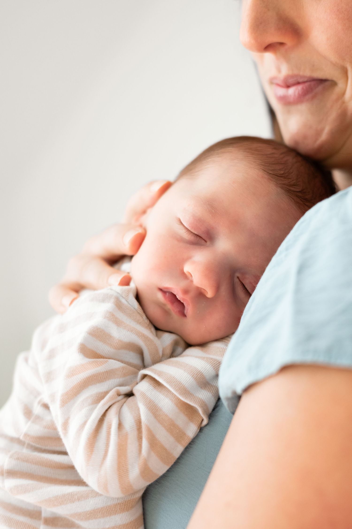 Newborn baby sleeping peacefully on mother's shoulder, wearing striped onesie.