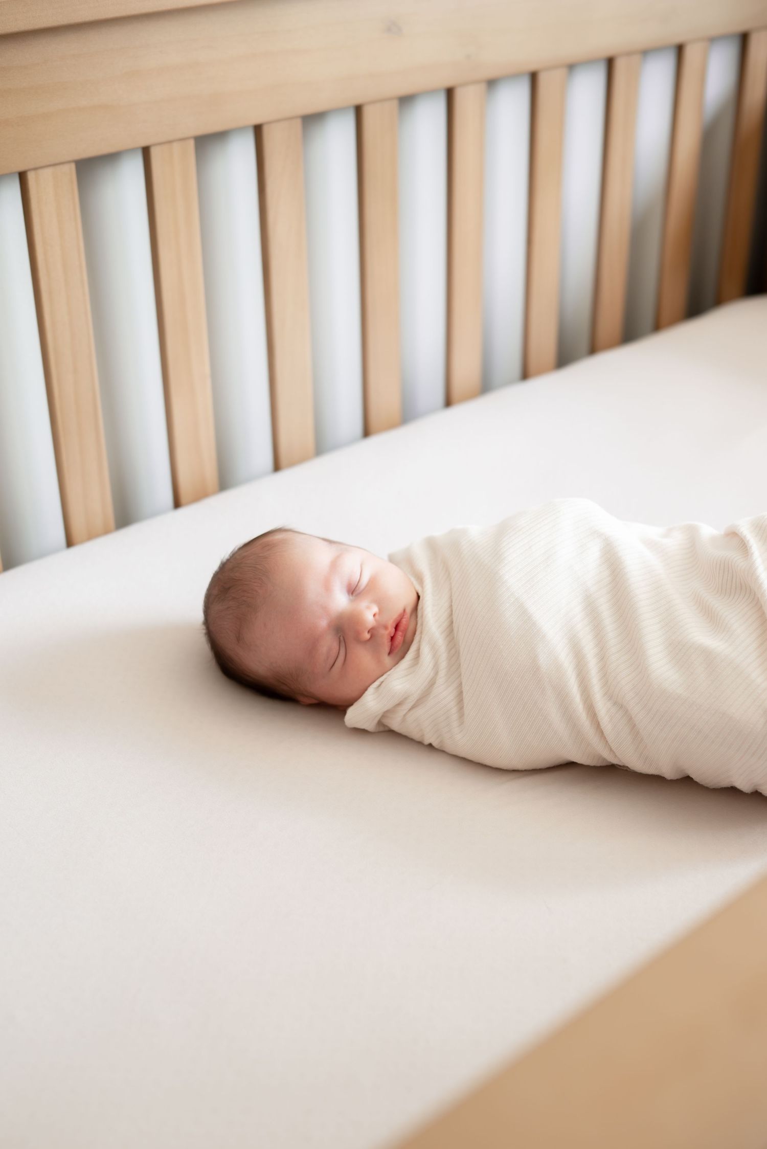 Newborn baby swaddled in white wrap sleeping peacefully in a wooden crib on a white fitted sheet.