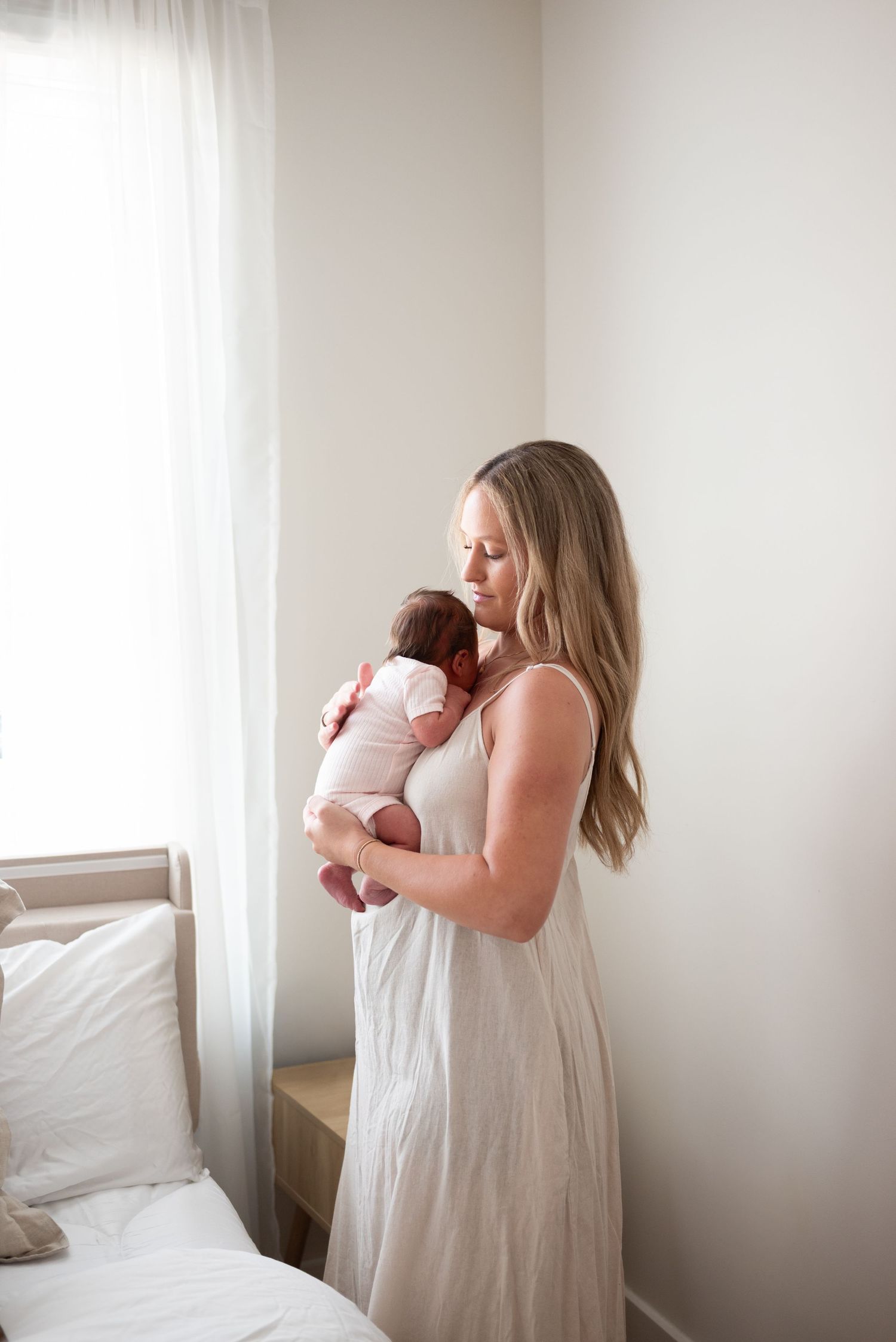 A mother in a white dress tenderly holds her newborn baby near a sunlit window in a serene bedroom.