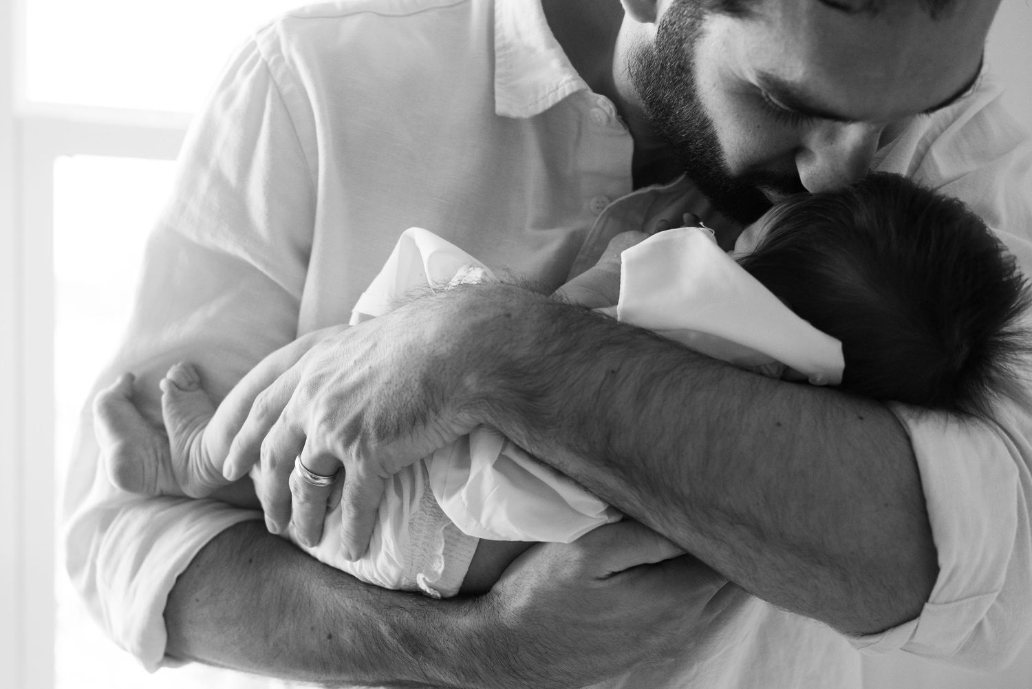 Black and white photo of a bearded father tenderly kissing his newborn baby while cradling it in his arms.
