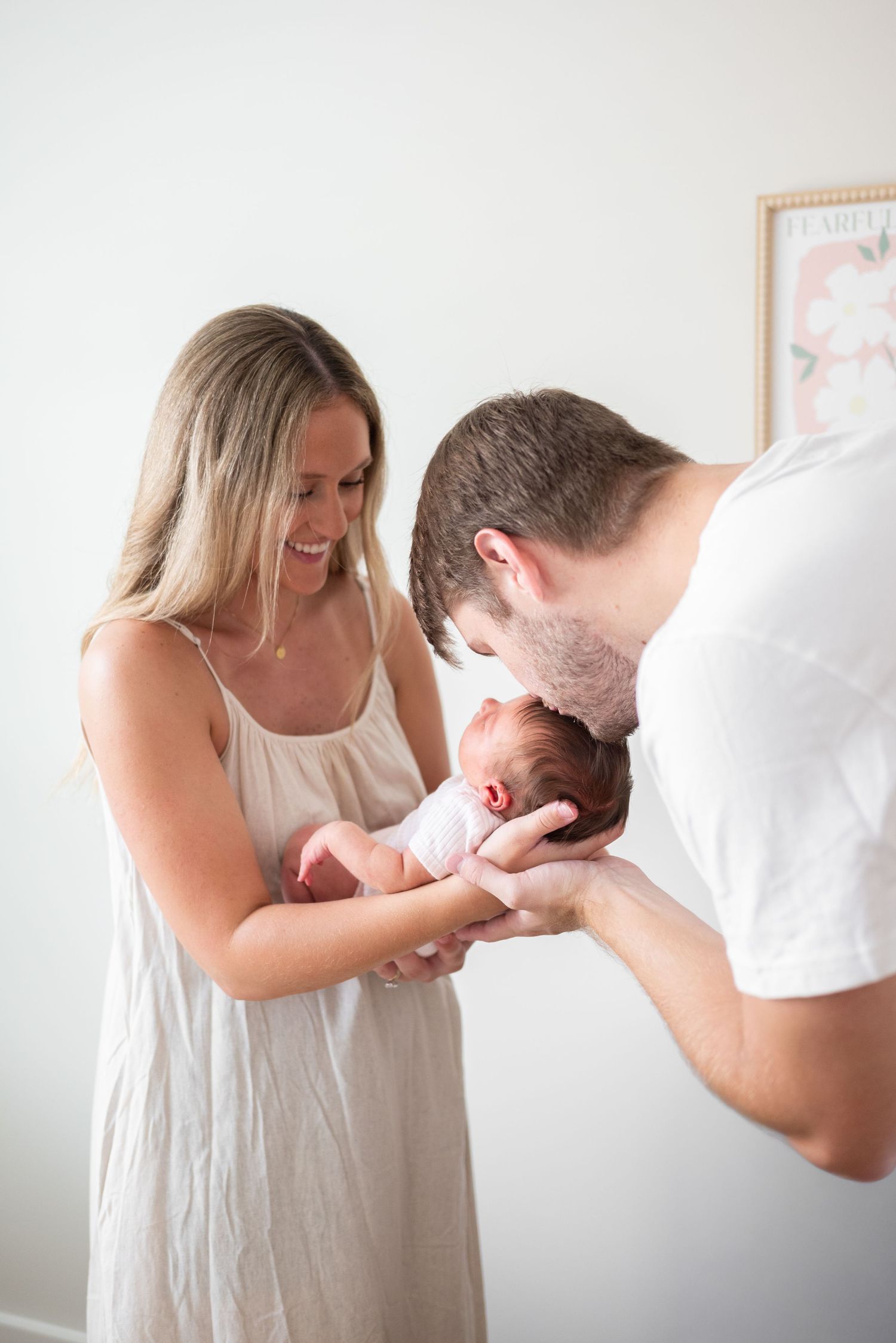 A smiling mother in a white dress holds her newborn baby while the father leans in to give the infant a gentle kiss.