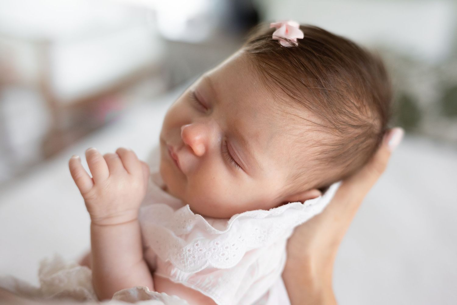 Peaceful newborn baby girl sleeping in white lace outfit with pink bow, cradled in loving arms.