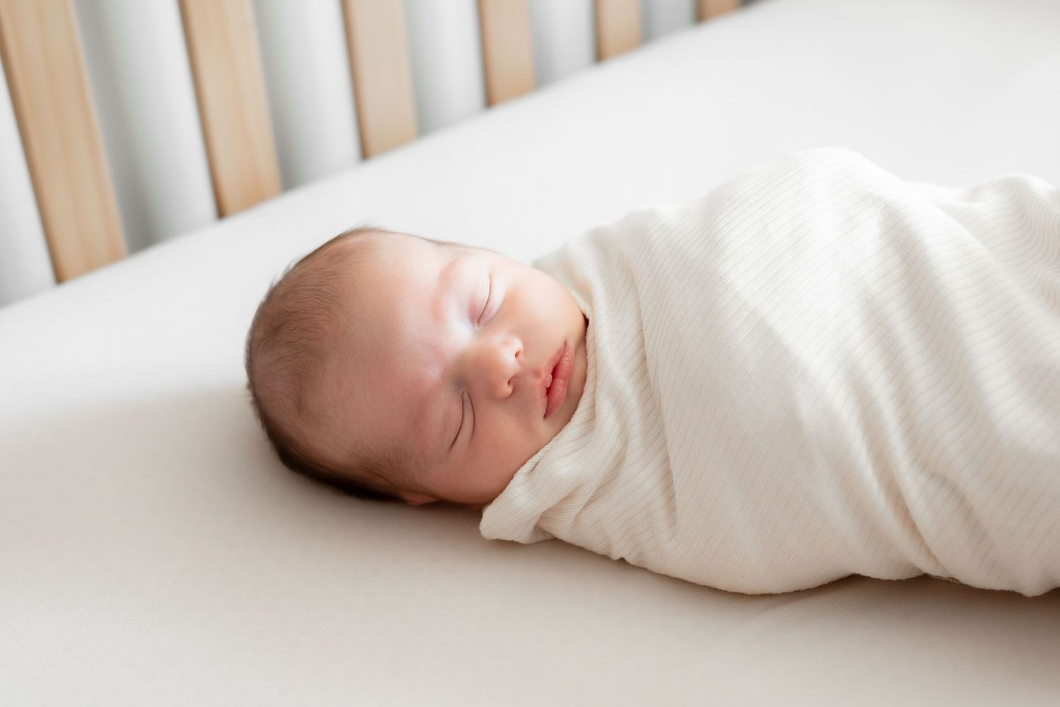 Swaddled newborn baby sleeping peacefully in a white crib, wrapped in a soft cream knit blanket.