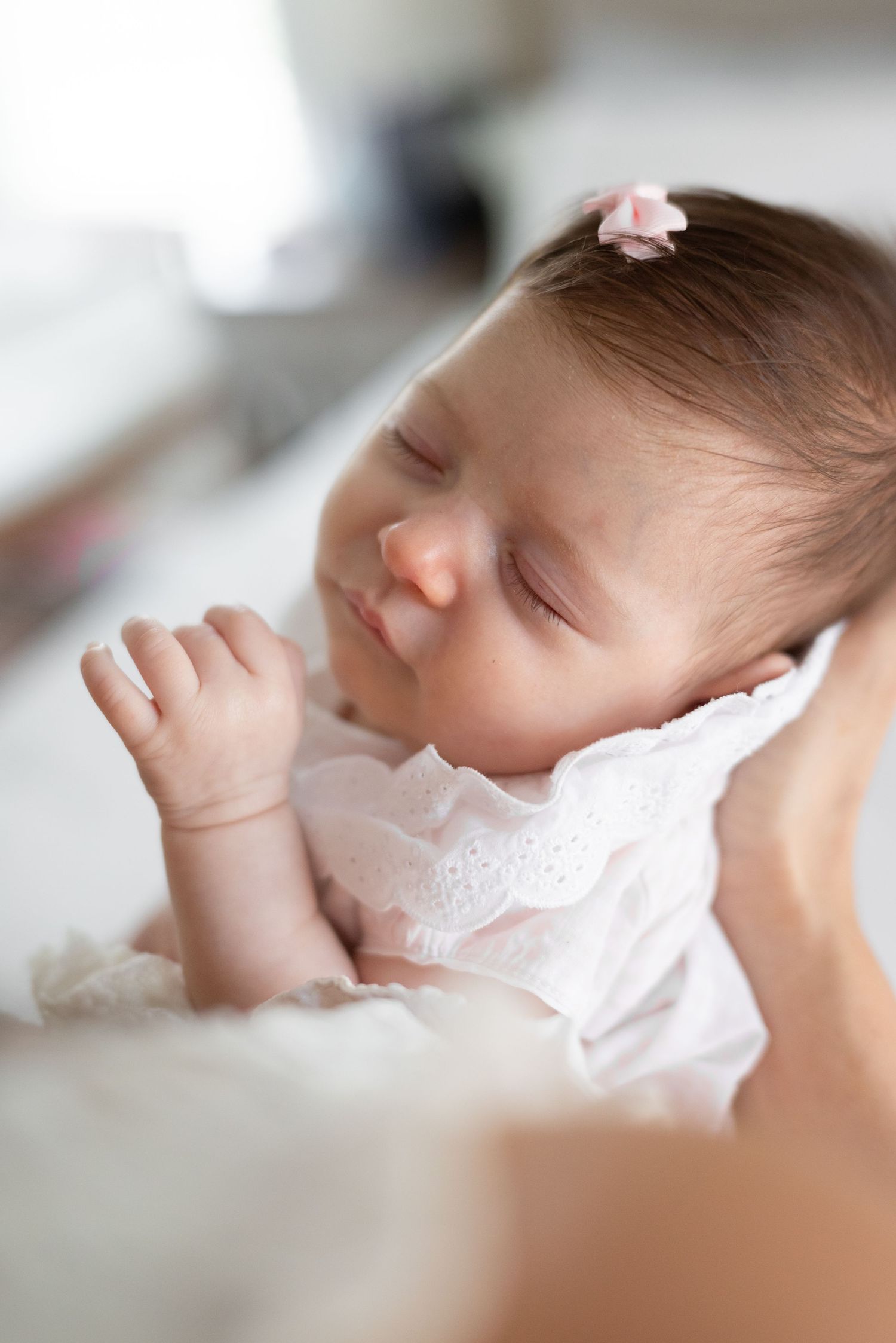Peaceful newborn baby girl sleeping in white lace outfit with pink bow, soft natural light portrait photography.
