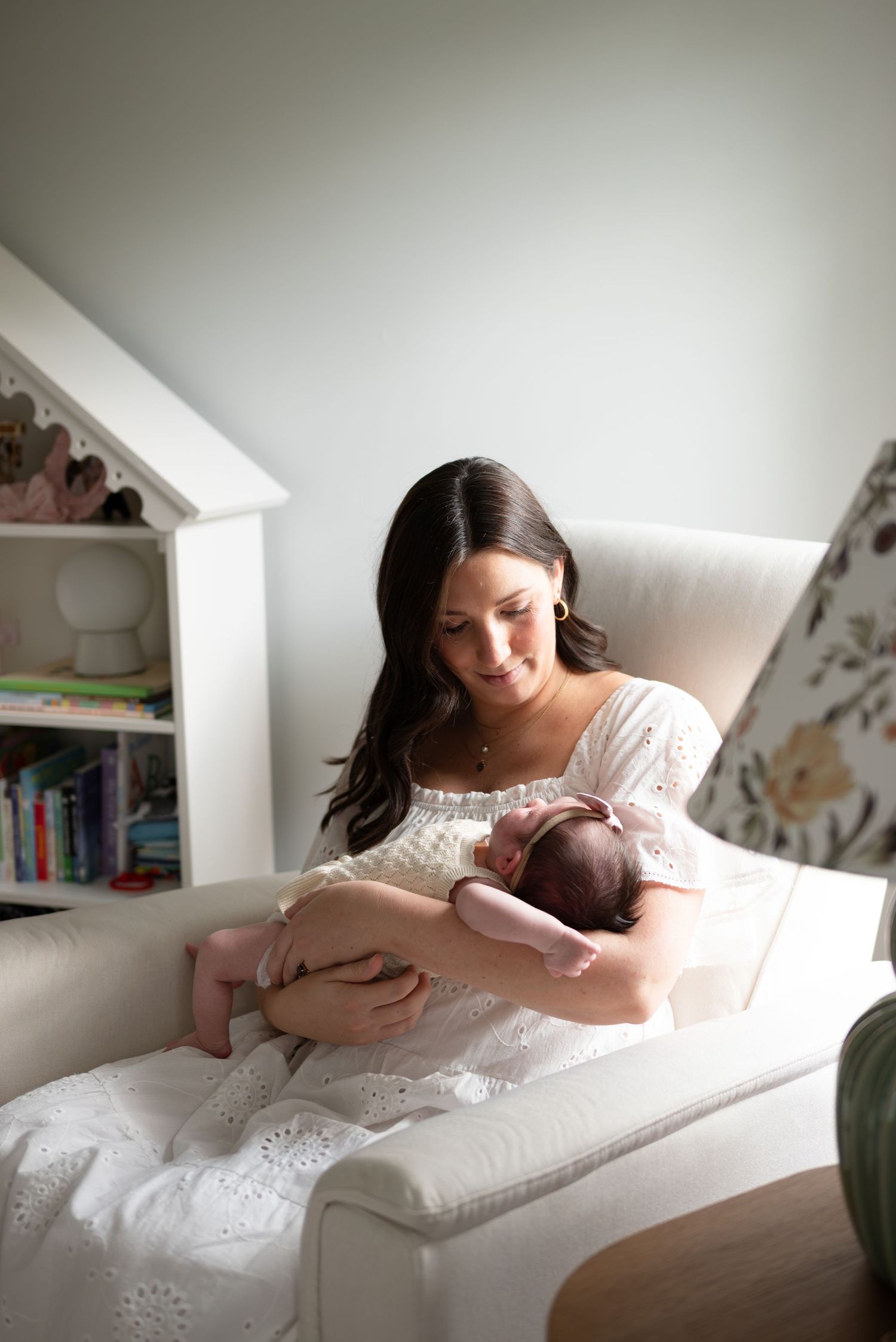 A mother in a white dress sits in a cream chair cradling her newborn baby in a softly decorated nursery.