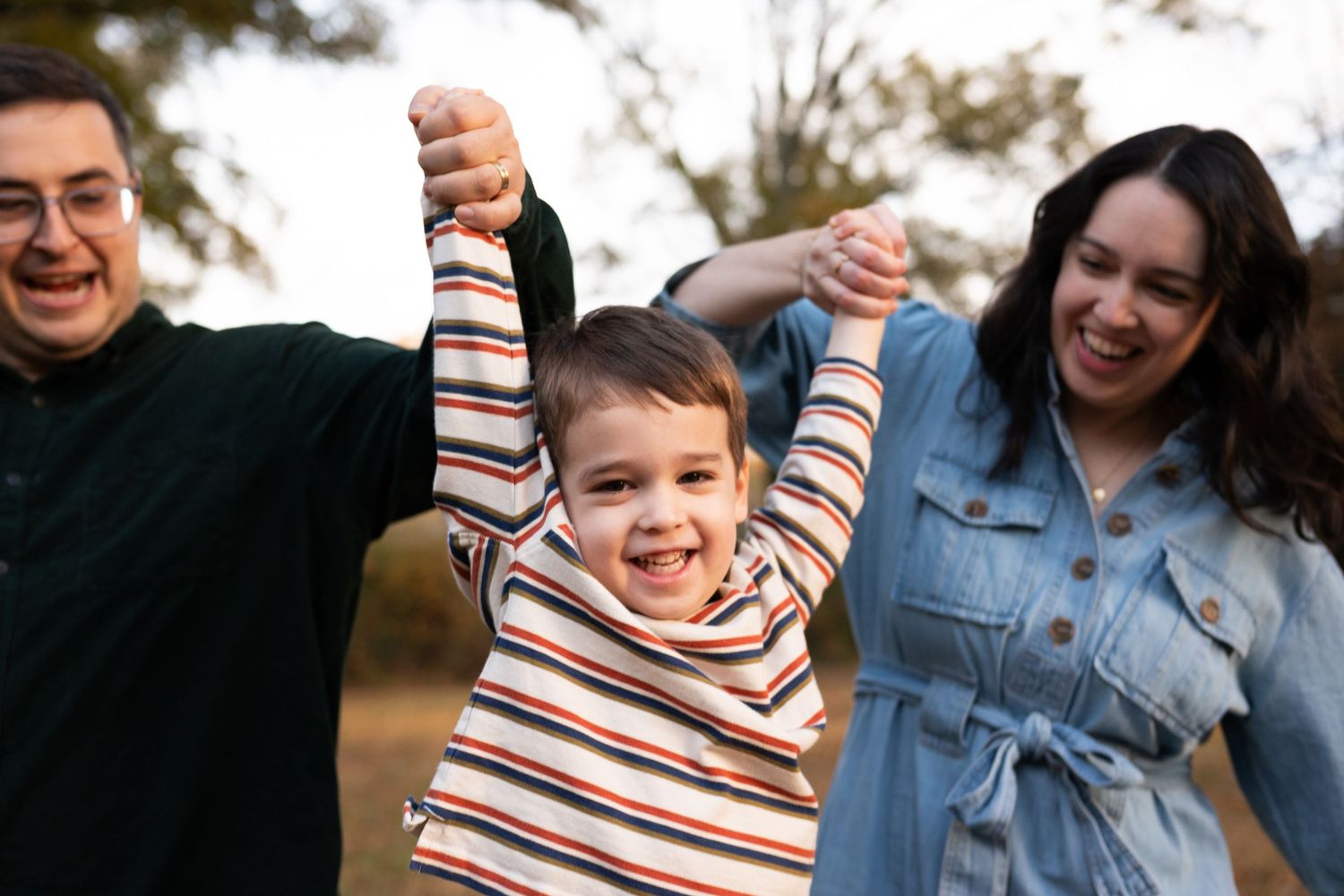 A joyful family of three holds hands outdoors, with a laughing boy being lifted by his smiling parents in a park.