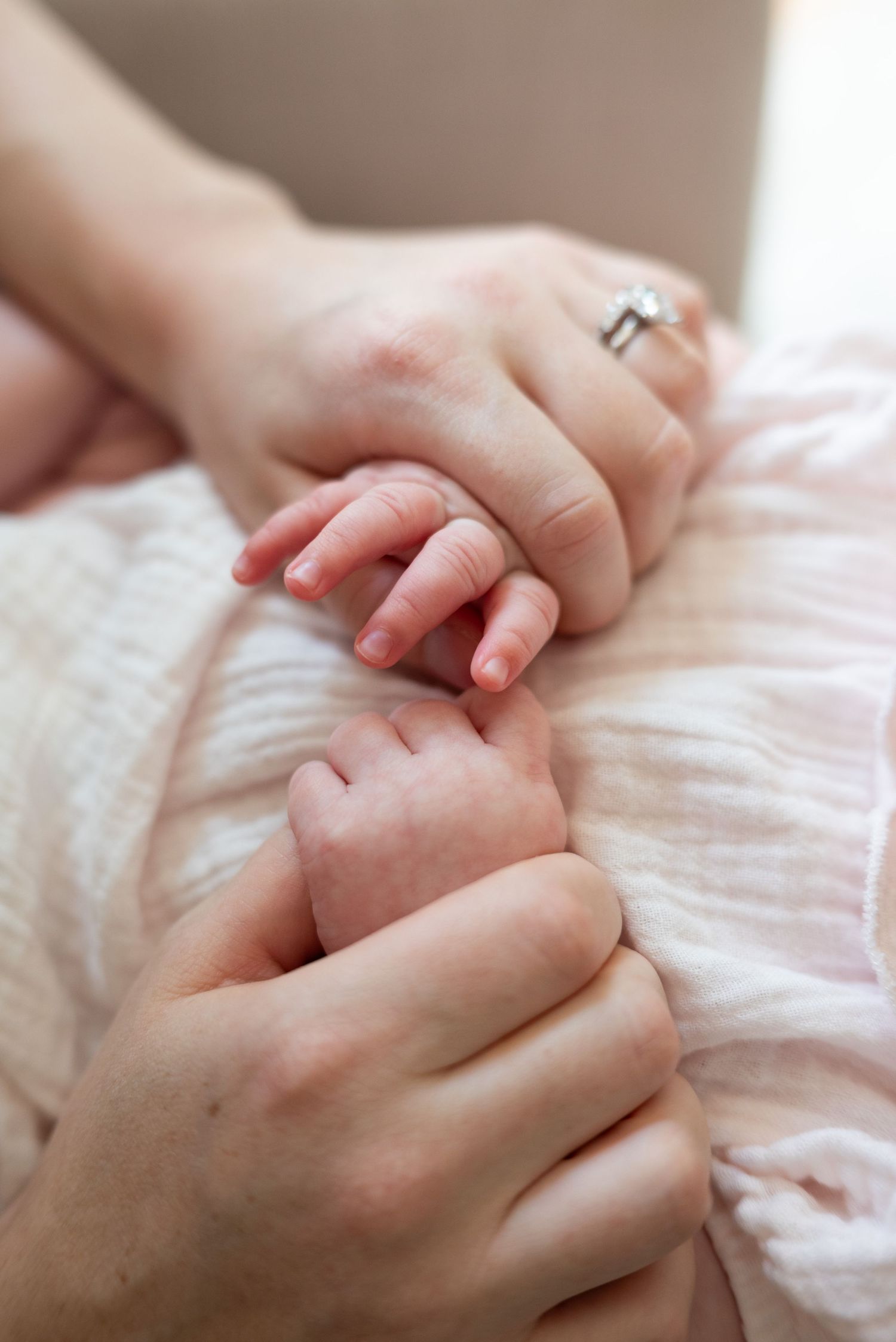 A mother gently holds her newborn baby's tiny hands, wearing a diamond ring, wrapped in a soft pink blanket.