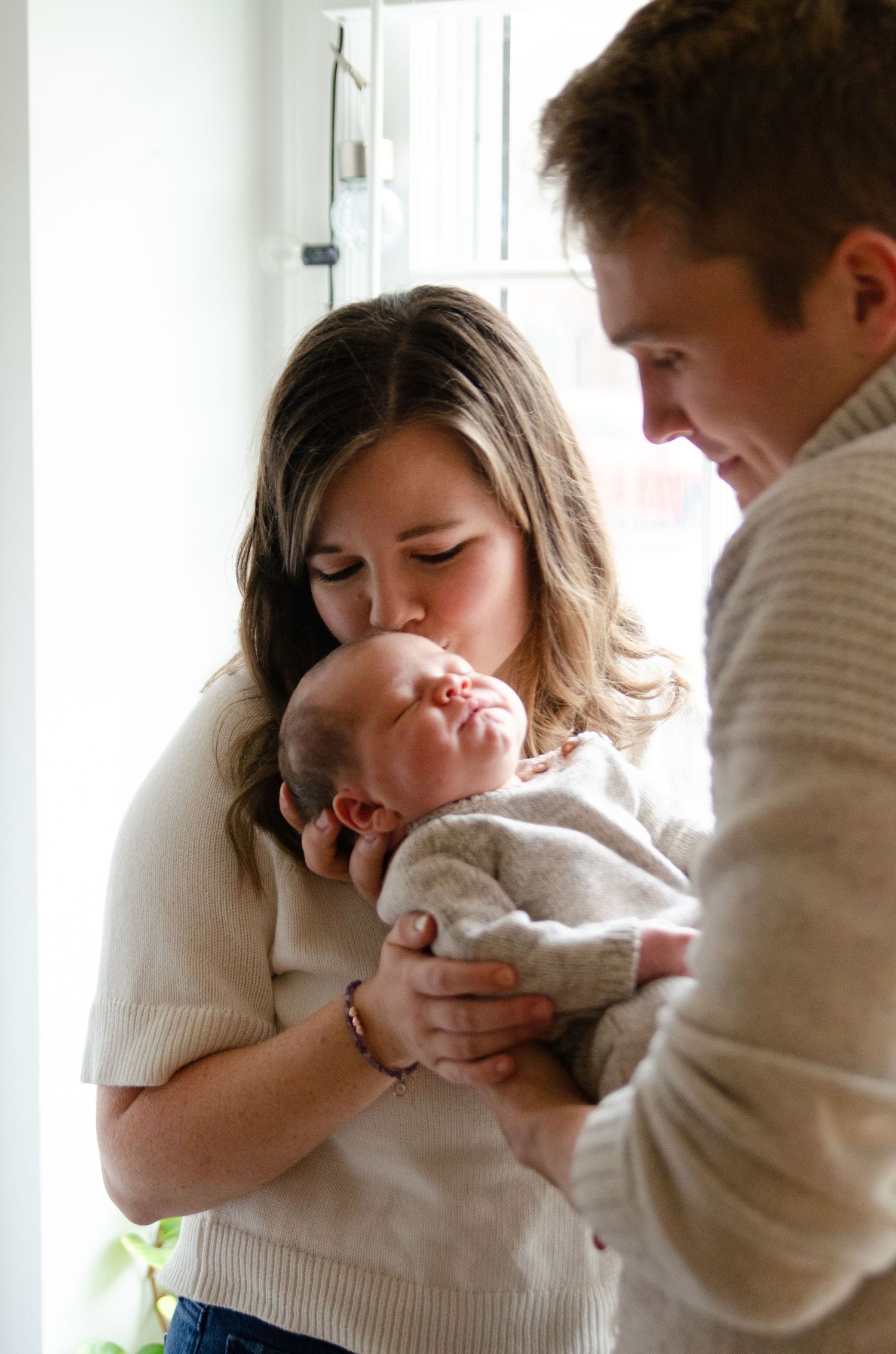 Family holding their newborn by a window while wearing cream sweaters during a lifestyle newborn photography session in Charlotte, NC.