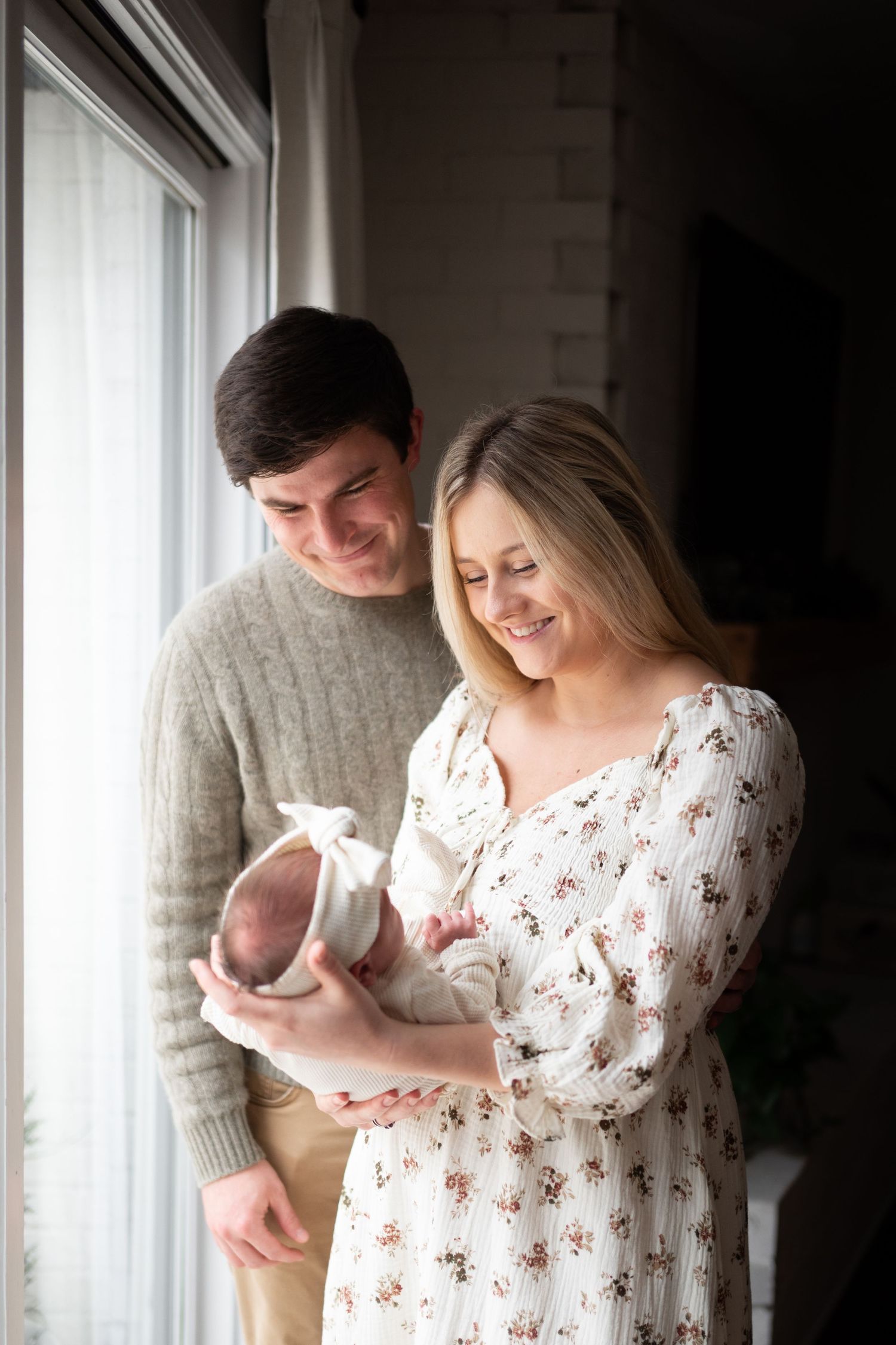 Happy couple smiling at their newborn baby wrapped in white near a bright window.