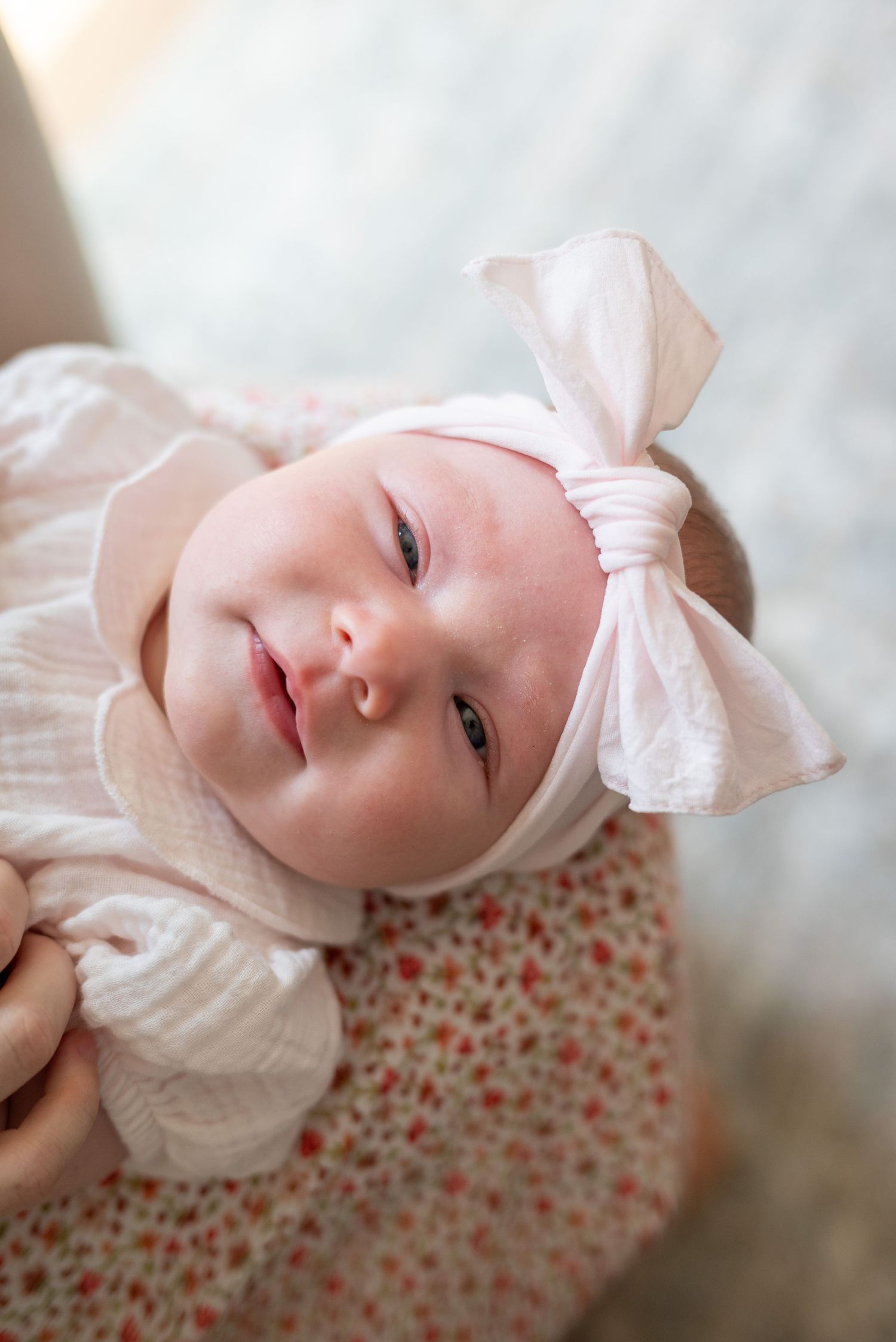 Newborn baby wearing a white bow headband and floral outfit, looking up with bright eyes and a gentle smile.