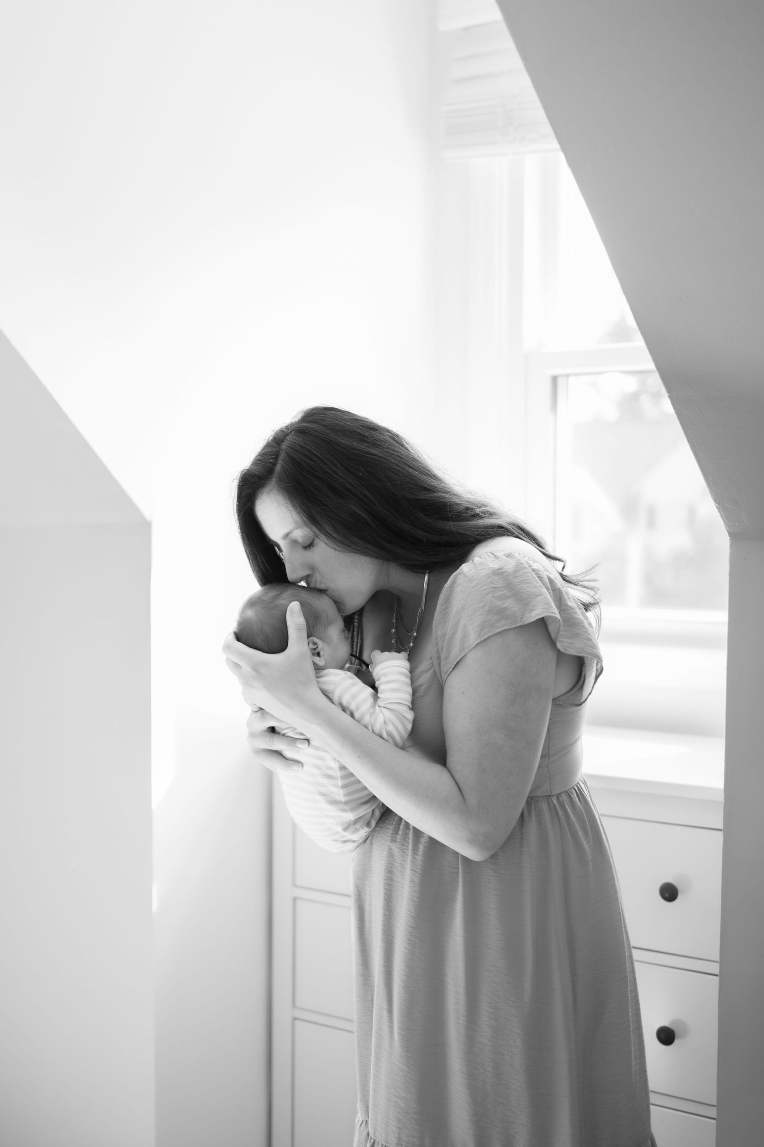 Black and white photo of a mother tenderly kissing her newborn baby near a bright window.