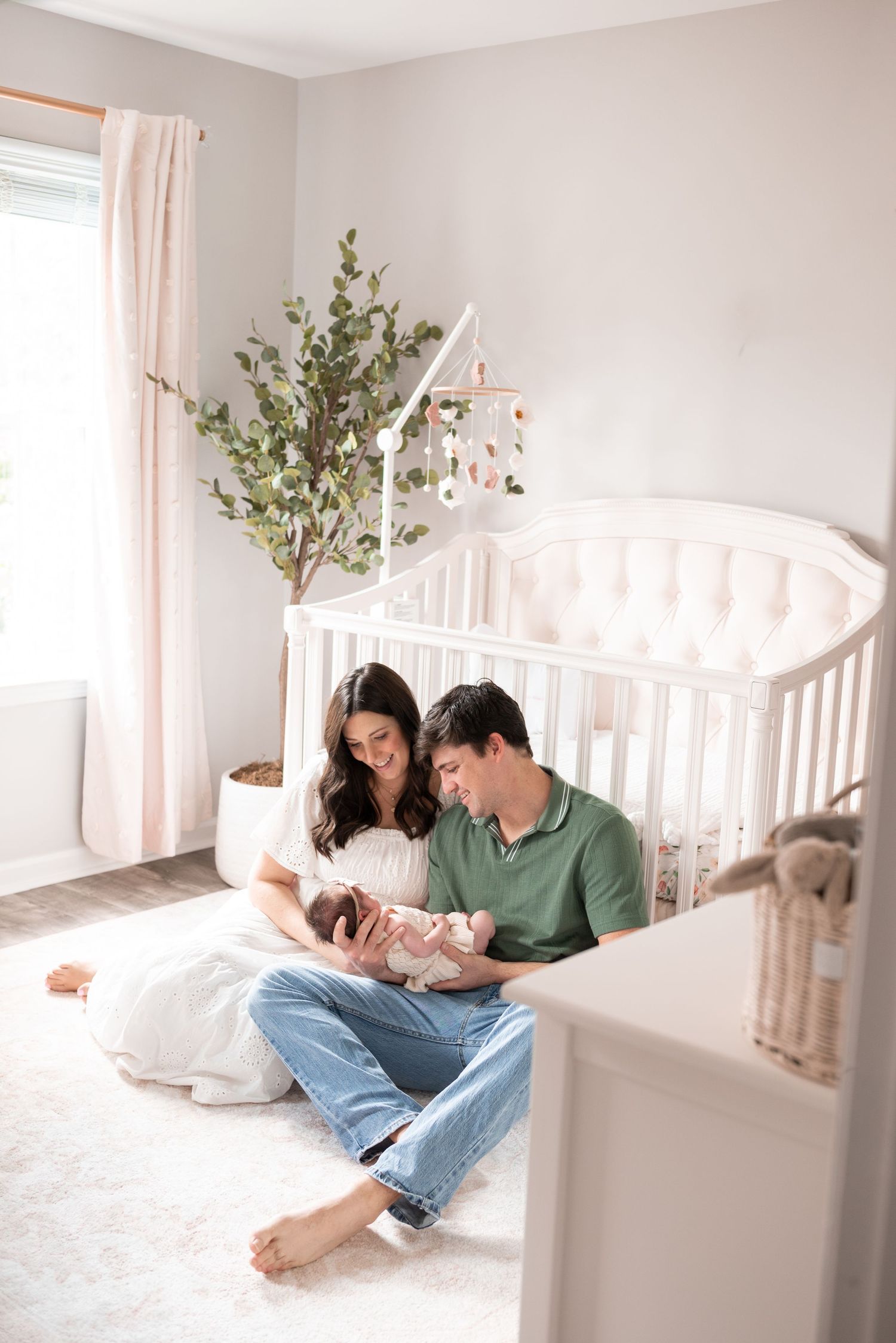 New parents sit on nursery floor holding newborn baby beside white oval crib with soft pink decor.