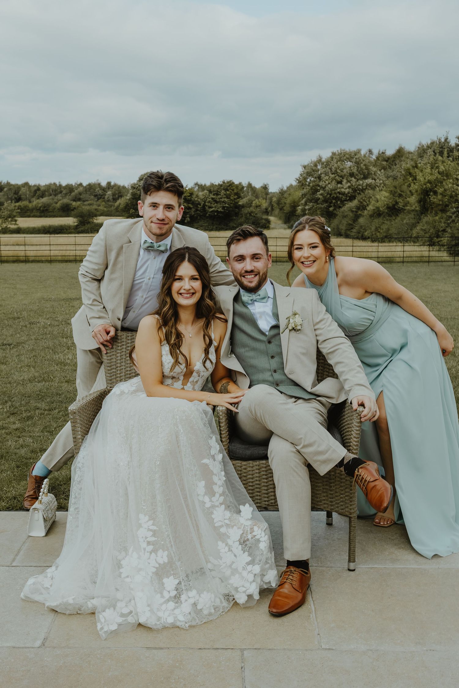Wedding party members in light colored attire pose together on outdoor patio with green field backdrop.