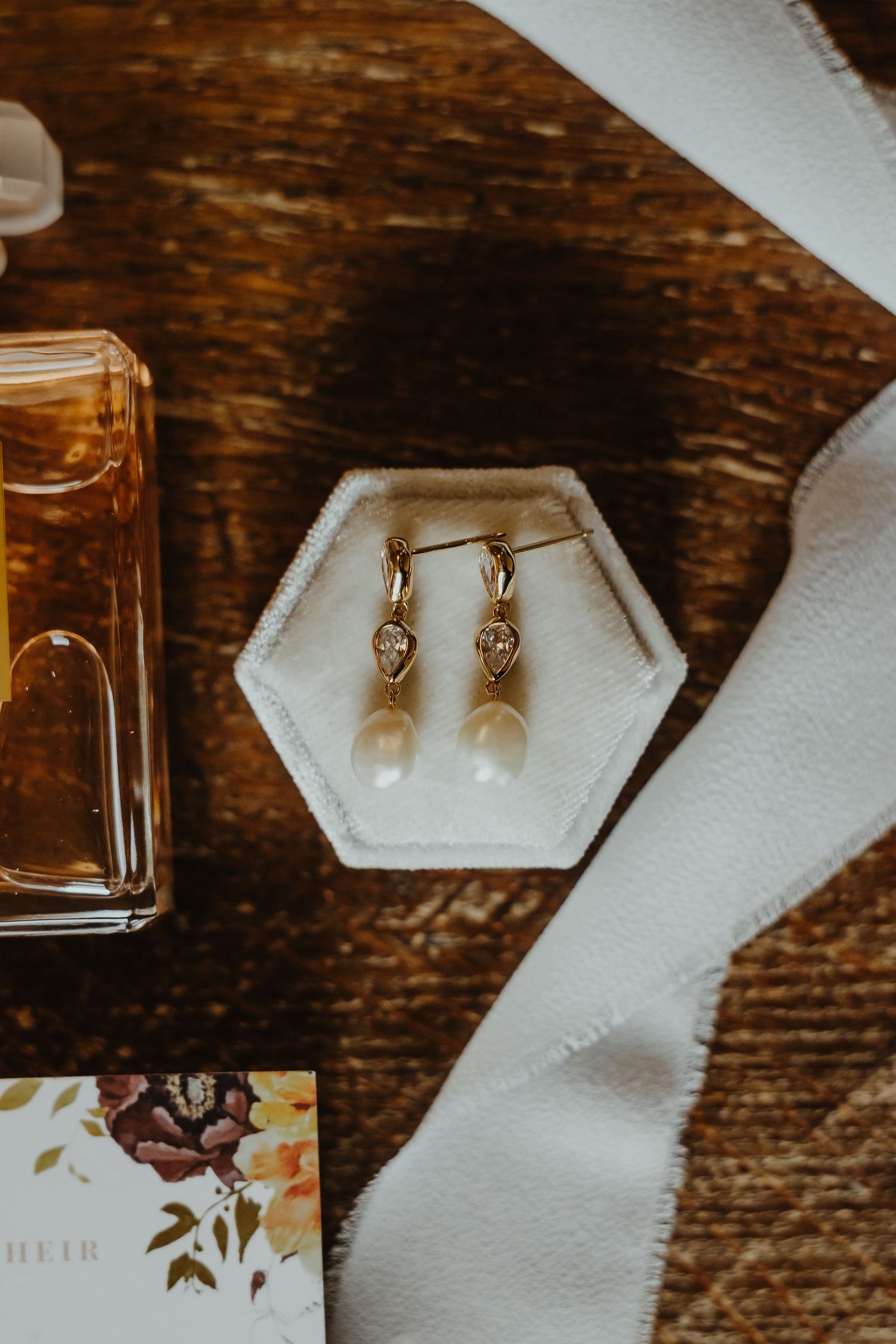 Pearl drop earrings and perfume bottle arranged on wooden surface.