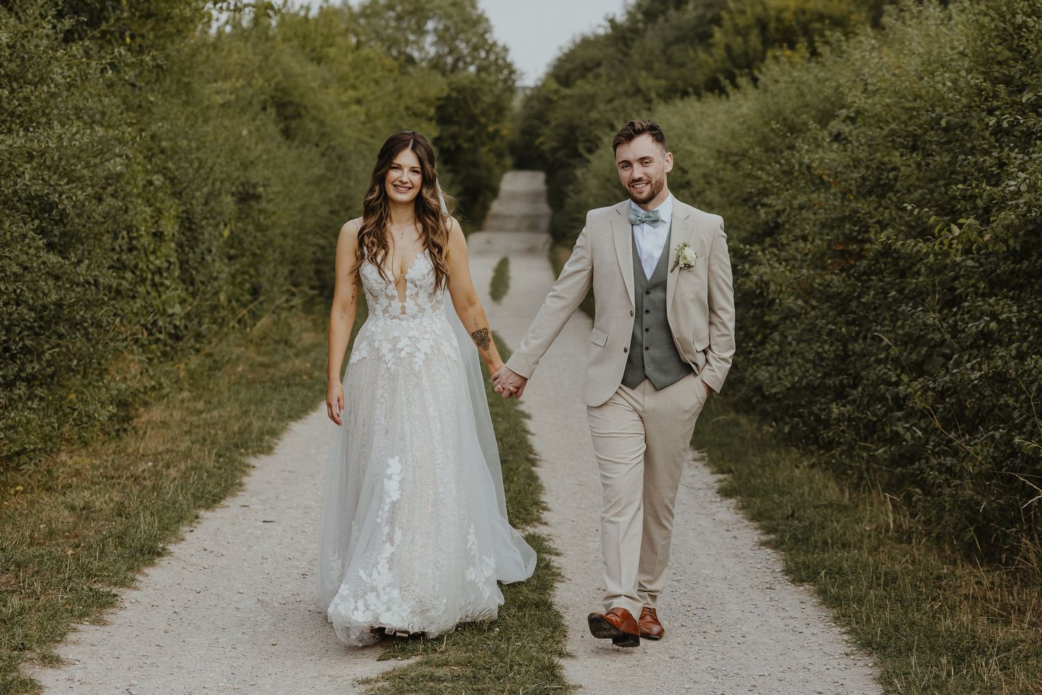 Wedding couple in light-colored attire walks along a scenic outdoor trail.