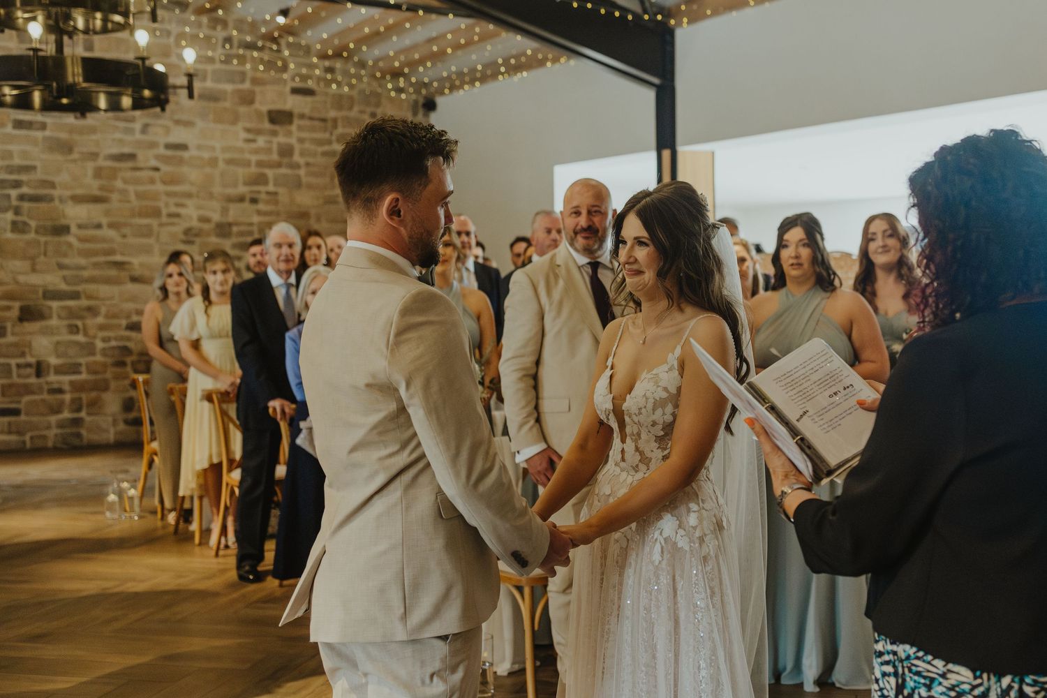 A couple exchanges vows during their wedding ceremony in a rustic brick-walled venue with guests looking on.