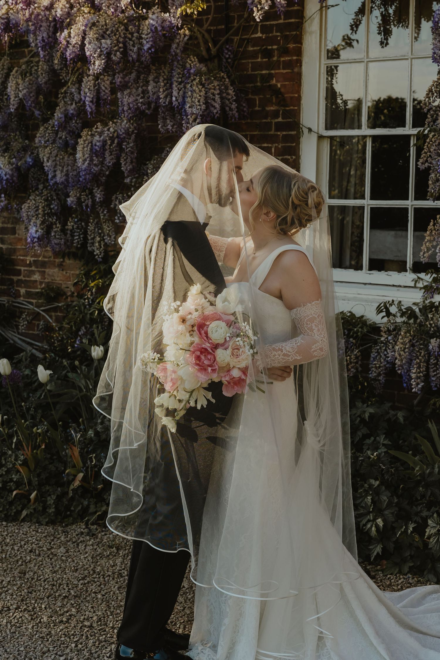 Bride and groom share a romantic kiss under a flowing veil, surrounded by wisteria blooms at an elegant wedding venue.