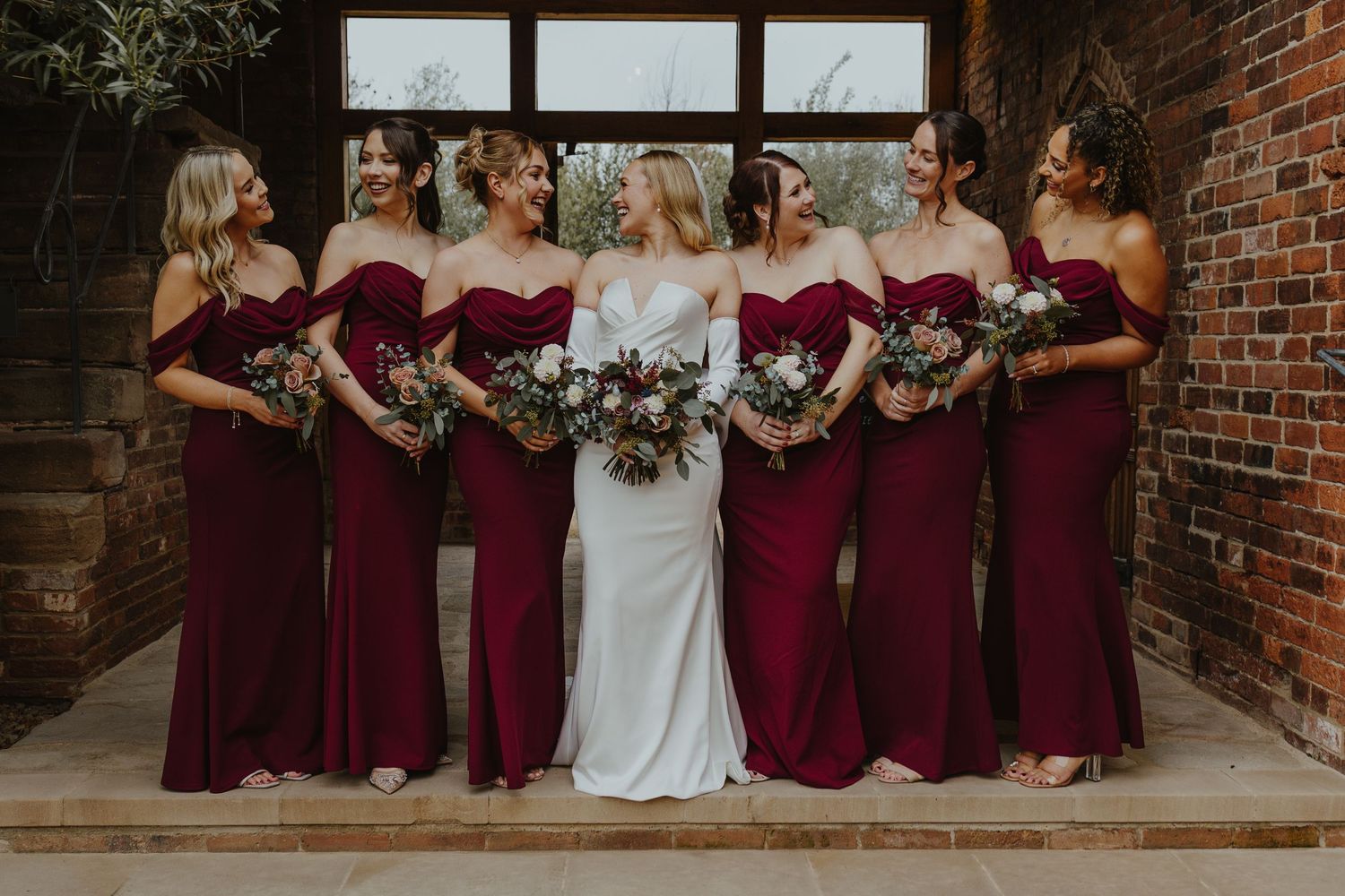 Bridesmaids in burgundy dresses pose with bride in white gown against brick wall.