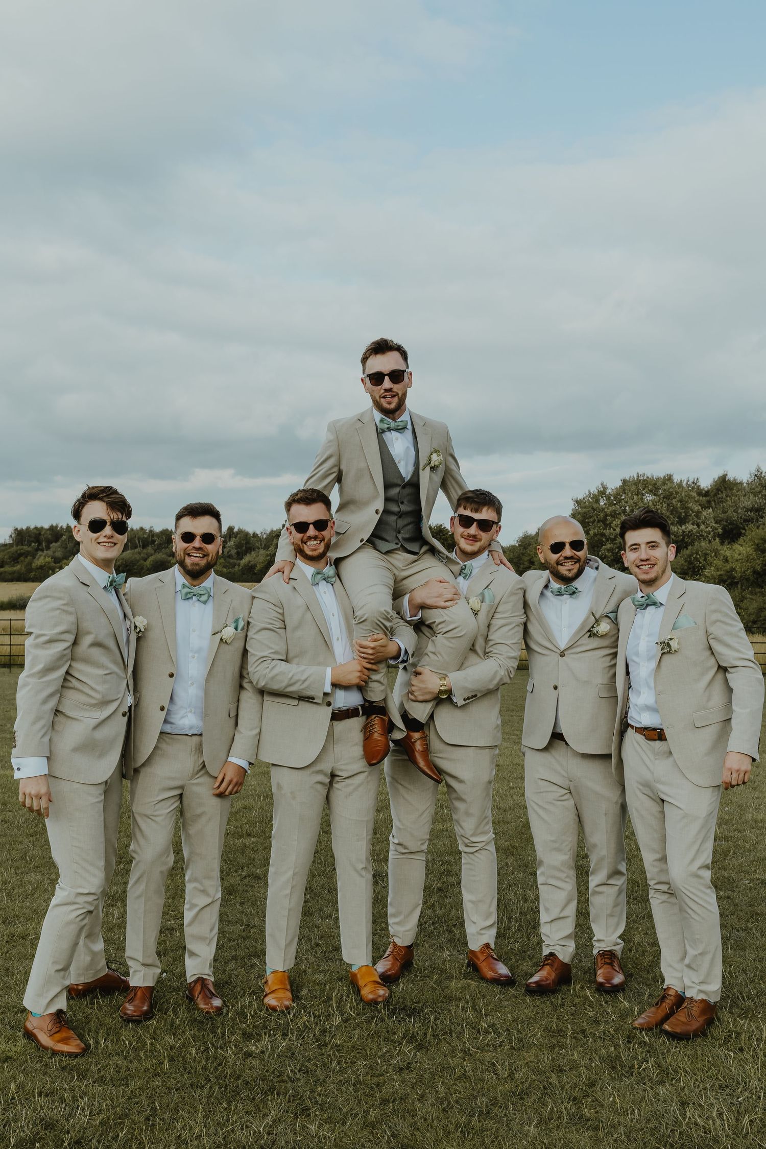Groomsmen in light gray suits lift groom in celebratory pose outdoors.