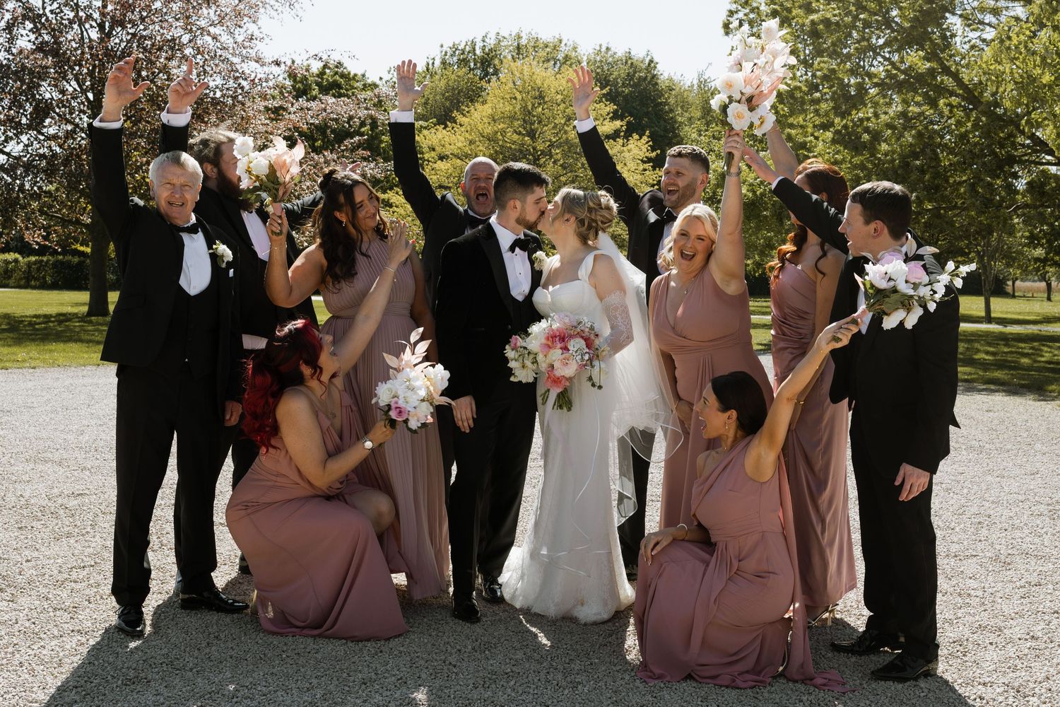 Wedding party celebrates outdoors, bridesmaids in mauve dresses, groomsmen in black suits, bride and groom kissing at center.