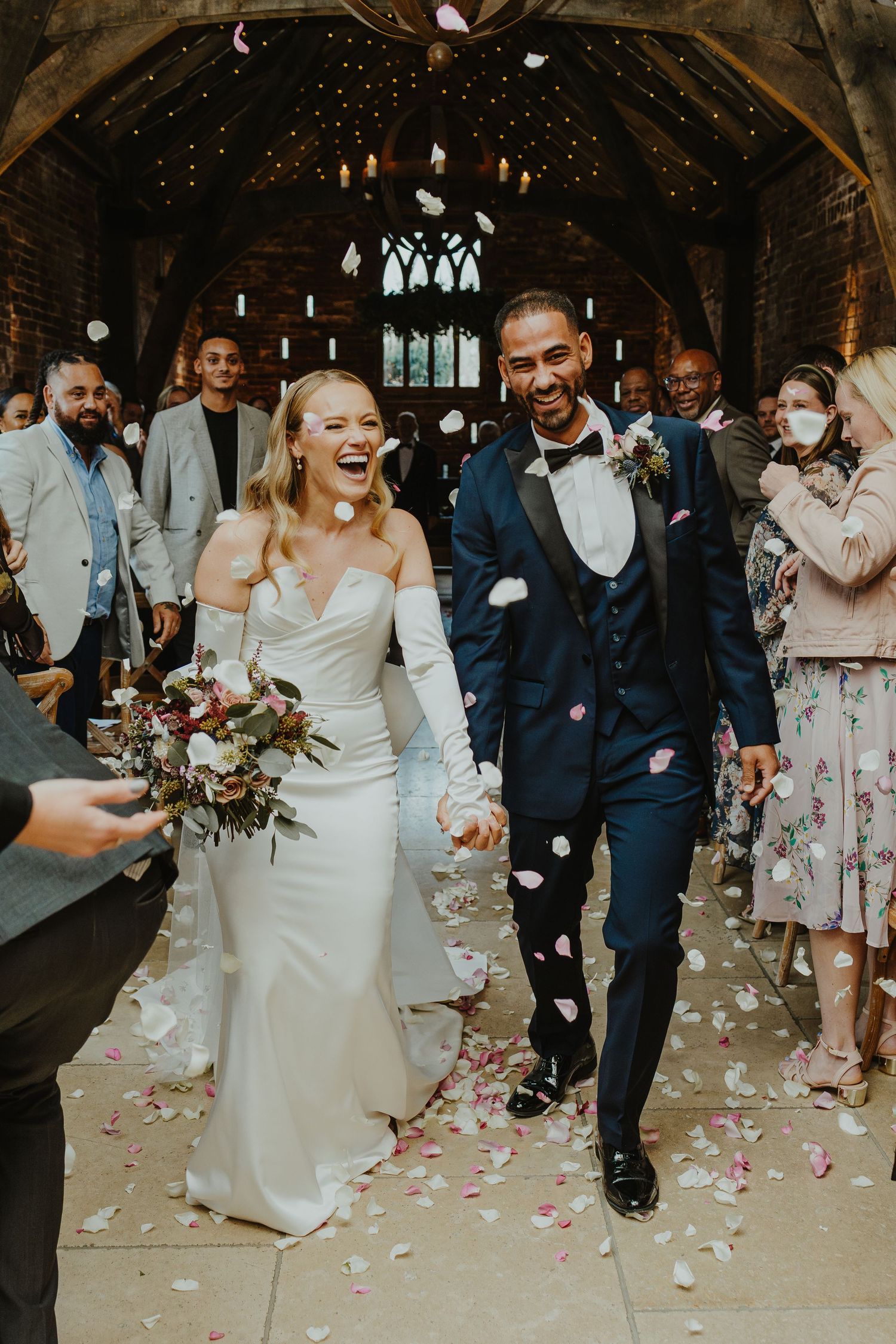 Newlyweds exit church ceremony under shower of pink rose petals with wedding guests cheering.