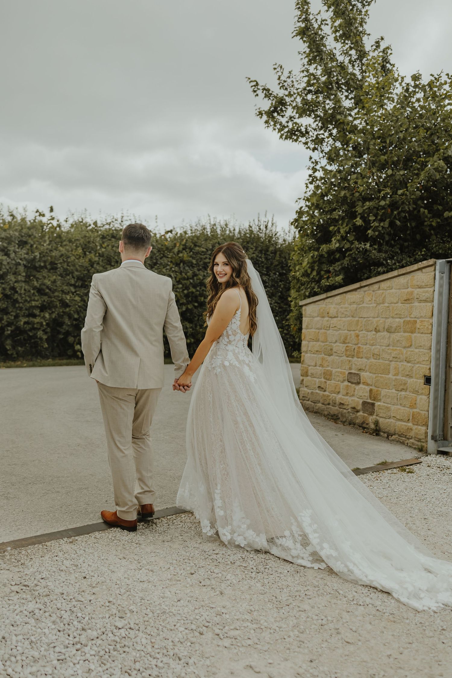 Bride and groom walking hand in hand on a gravel path with stone wall backdrop.