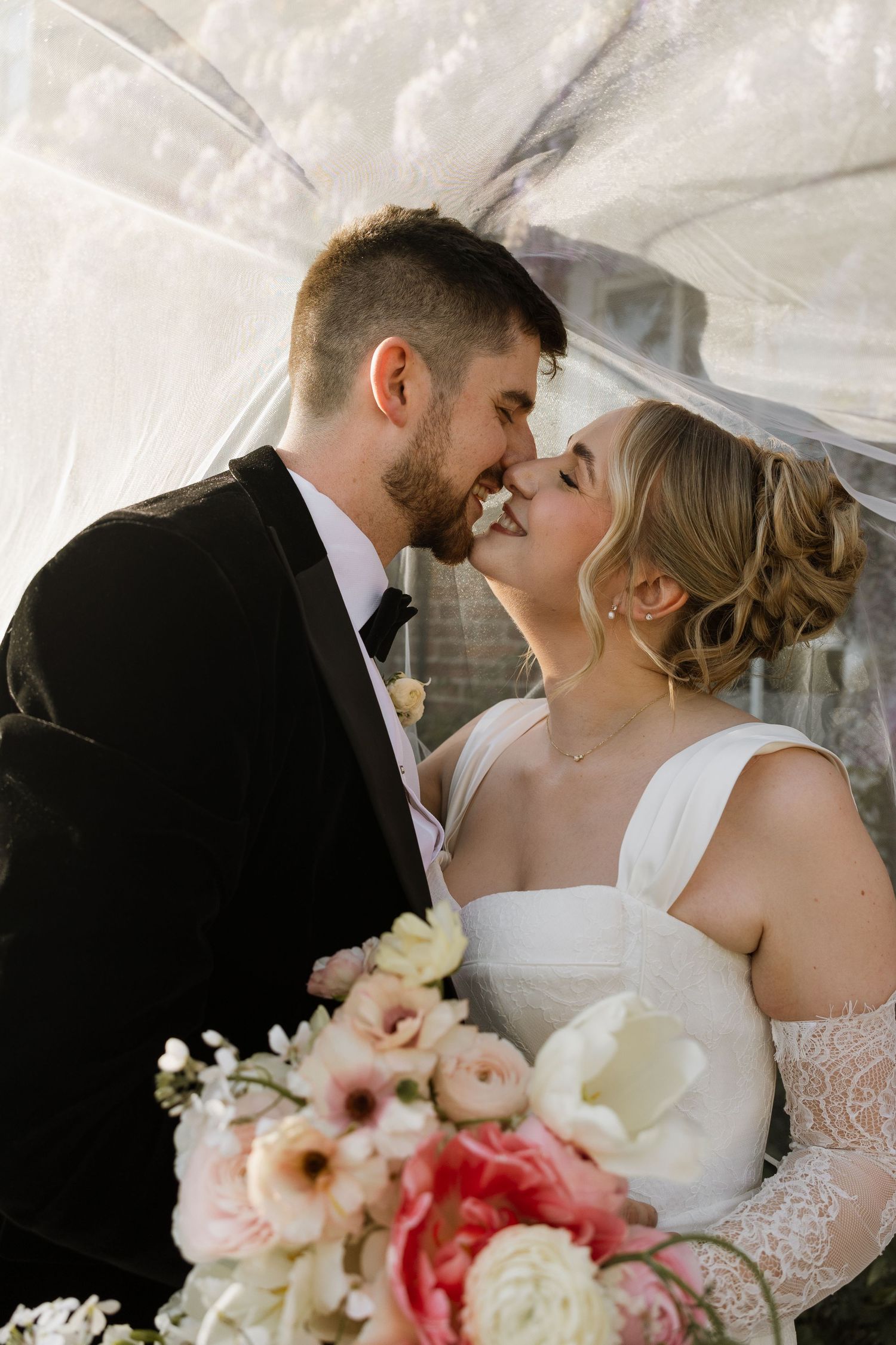 Bride and groom sharing a romantic moment under a veil, with a pink and white floral bouquet in the foreground.