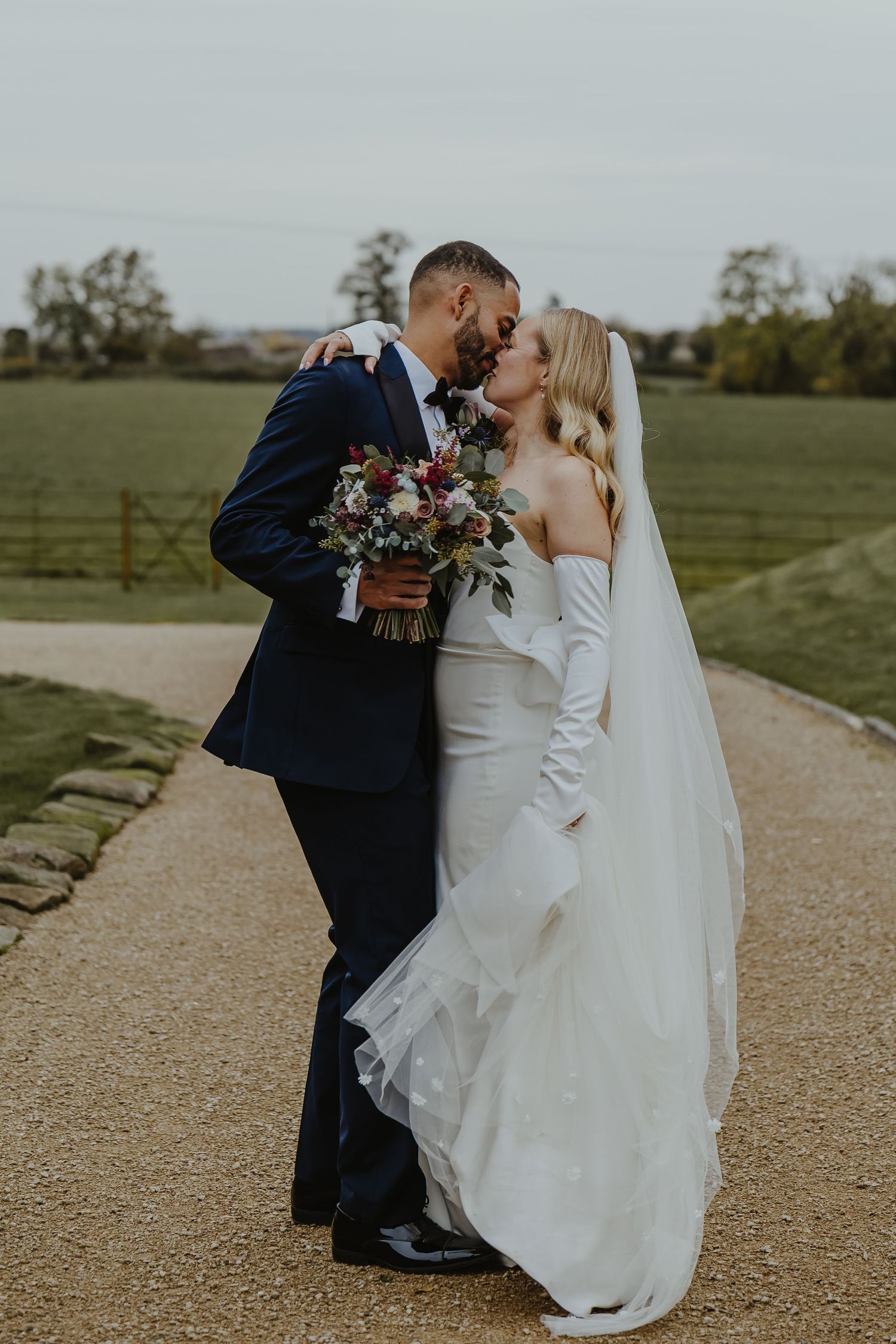 Newlyweds share a romantic kiss on a country path while holding a floral bouquet.