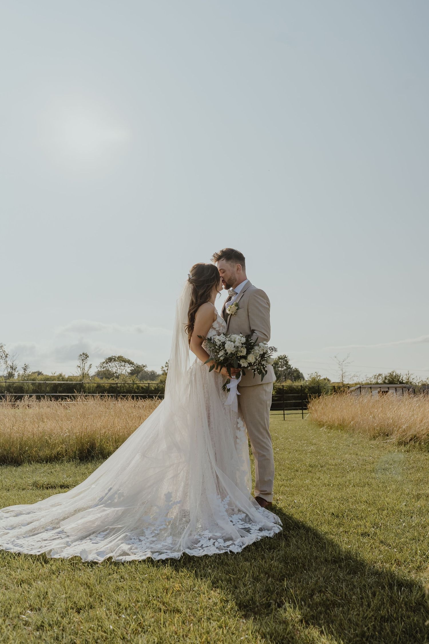 Elegant wedding couple embracing in field with bride's lace train flowing behind.