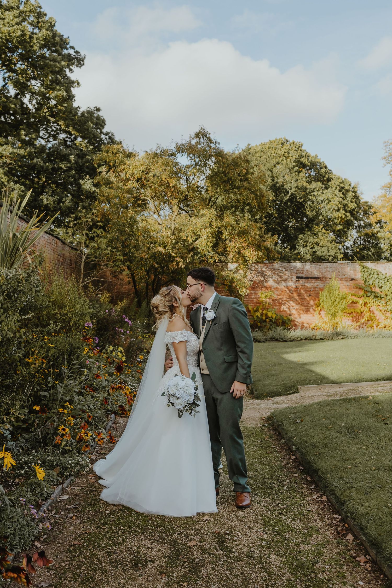 Newlyweds walking along garden path surrounded by autumn foliage.