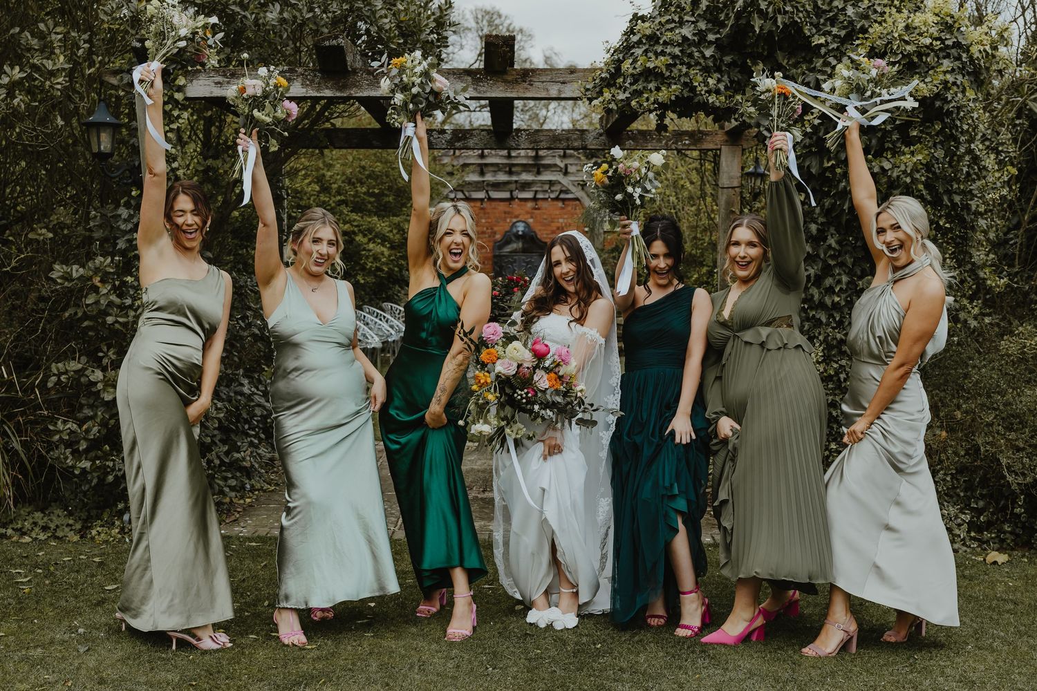 Bride in white gown with six bridesmaids in green and sage dresses celebrating outdoors, raising arms joyfully.