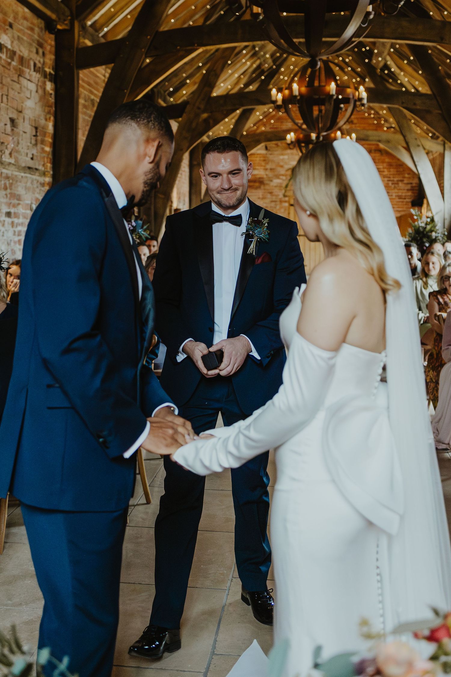 A wedding ceremony in progress inside a rustic venue with exposed wooden beams and twinkle lights.