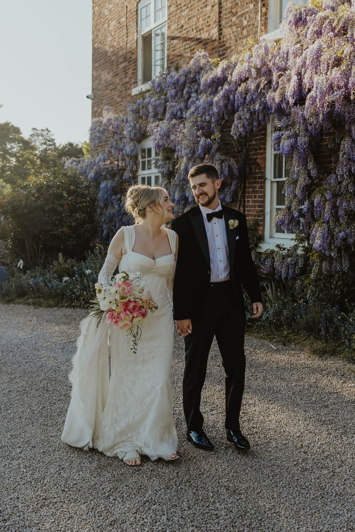 Bride and groom walk hand in hand before a wisteria-covered brick manor, bride holding a pink bouquet in golden light.