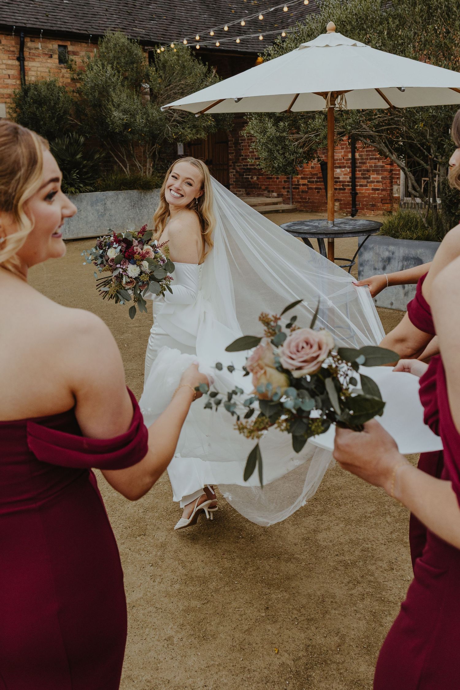 Wedding party playfully poses with bride's veil in outdoor garden setting.