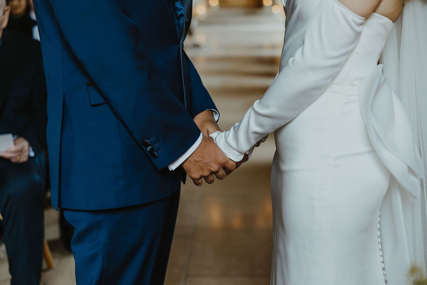 Close-up of joined hands between a couple in wedding attire showing blue suit sleeve and white dress details.